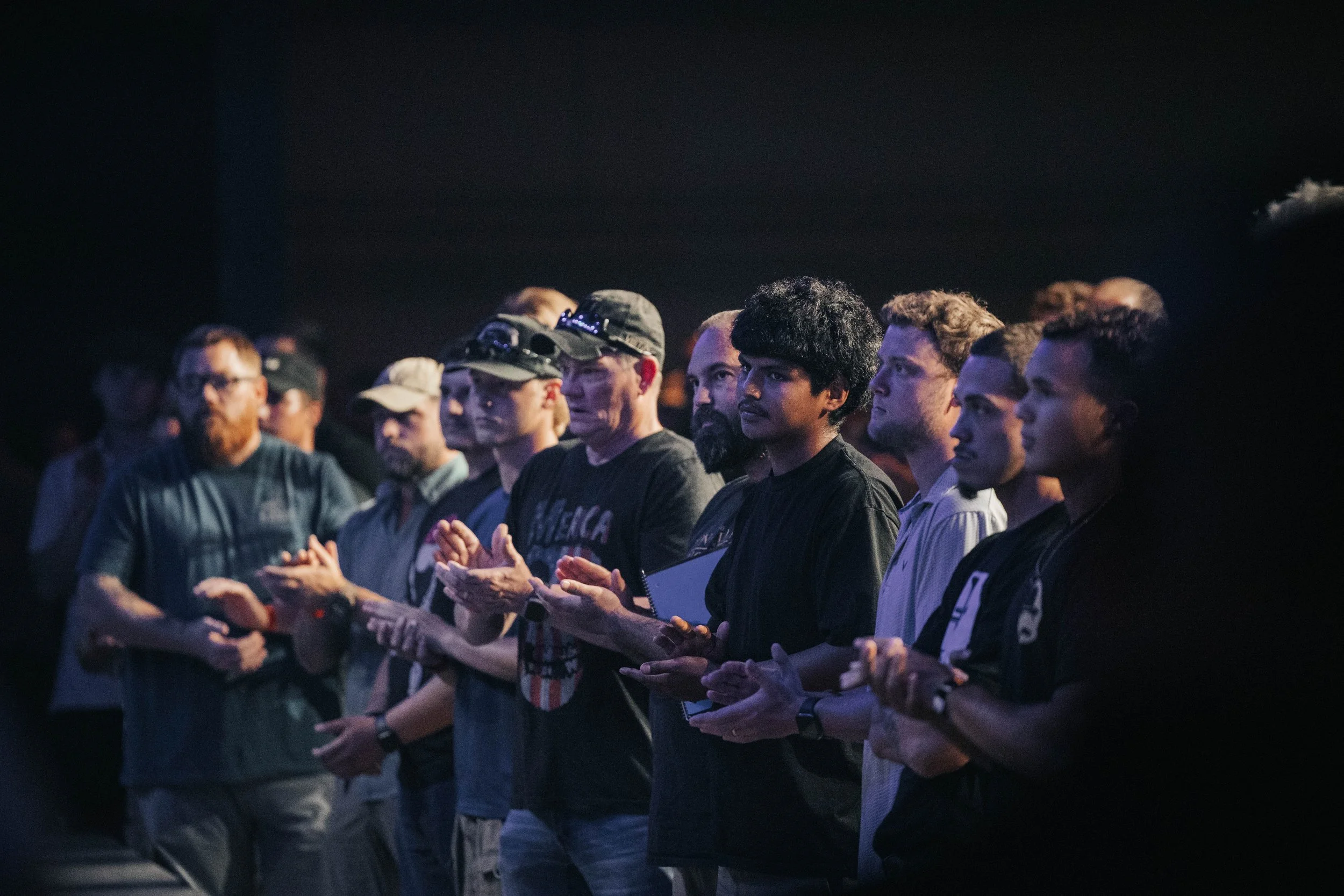 Group of people standing and clapping at an event, facing toward a stage or speaker, in a dimly lit environment.