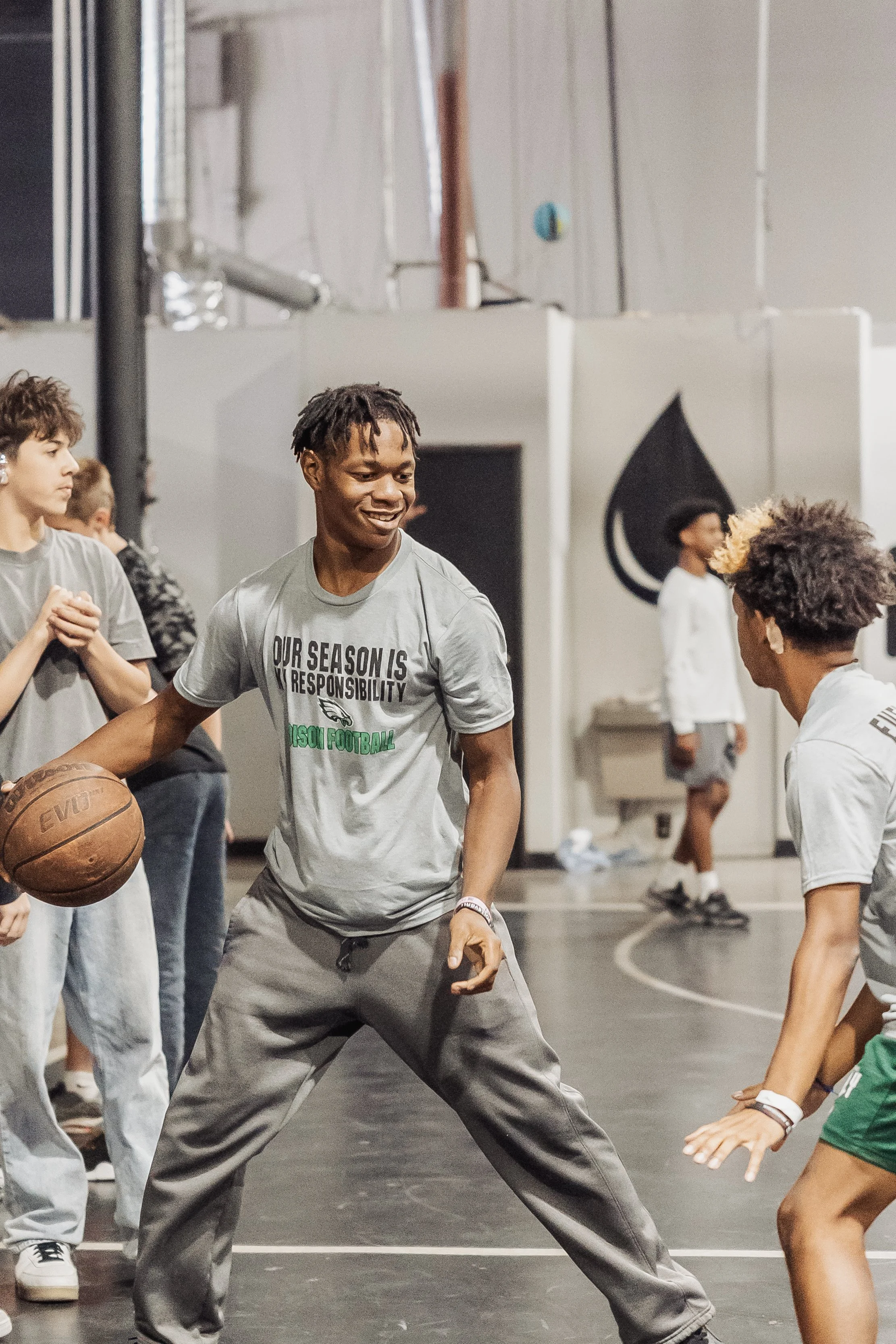 Young men and boys in workout clothes practicing basketball on an indoor court, with some wearing T-shirts with messages, and one smiling while holding a basketball.
