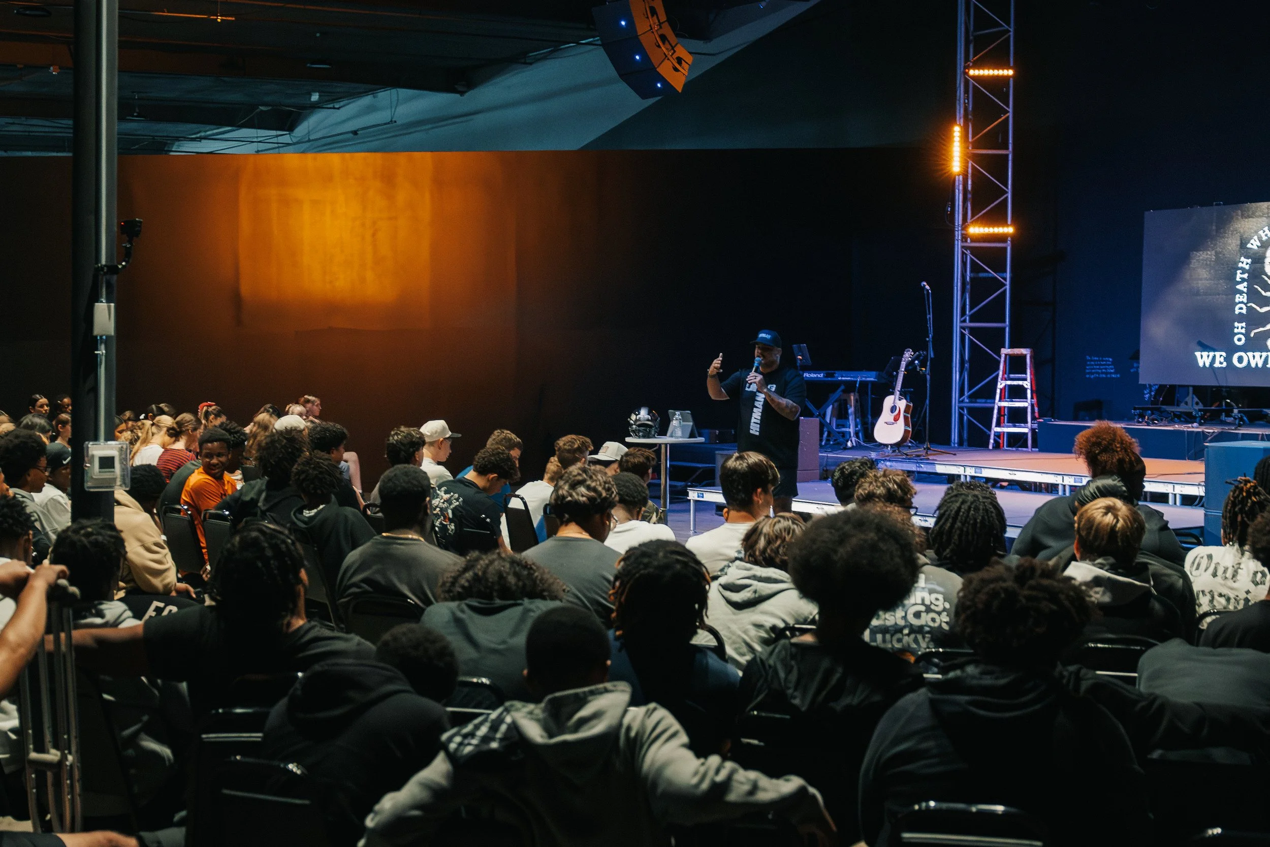 A speaker presenting on stage at a large indoor event with a seated audience. The stage has musical instruments and a large screen displaying the text "WE OW" and part of a larger message. The event appears to be a religious or motivational gathering