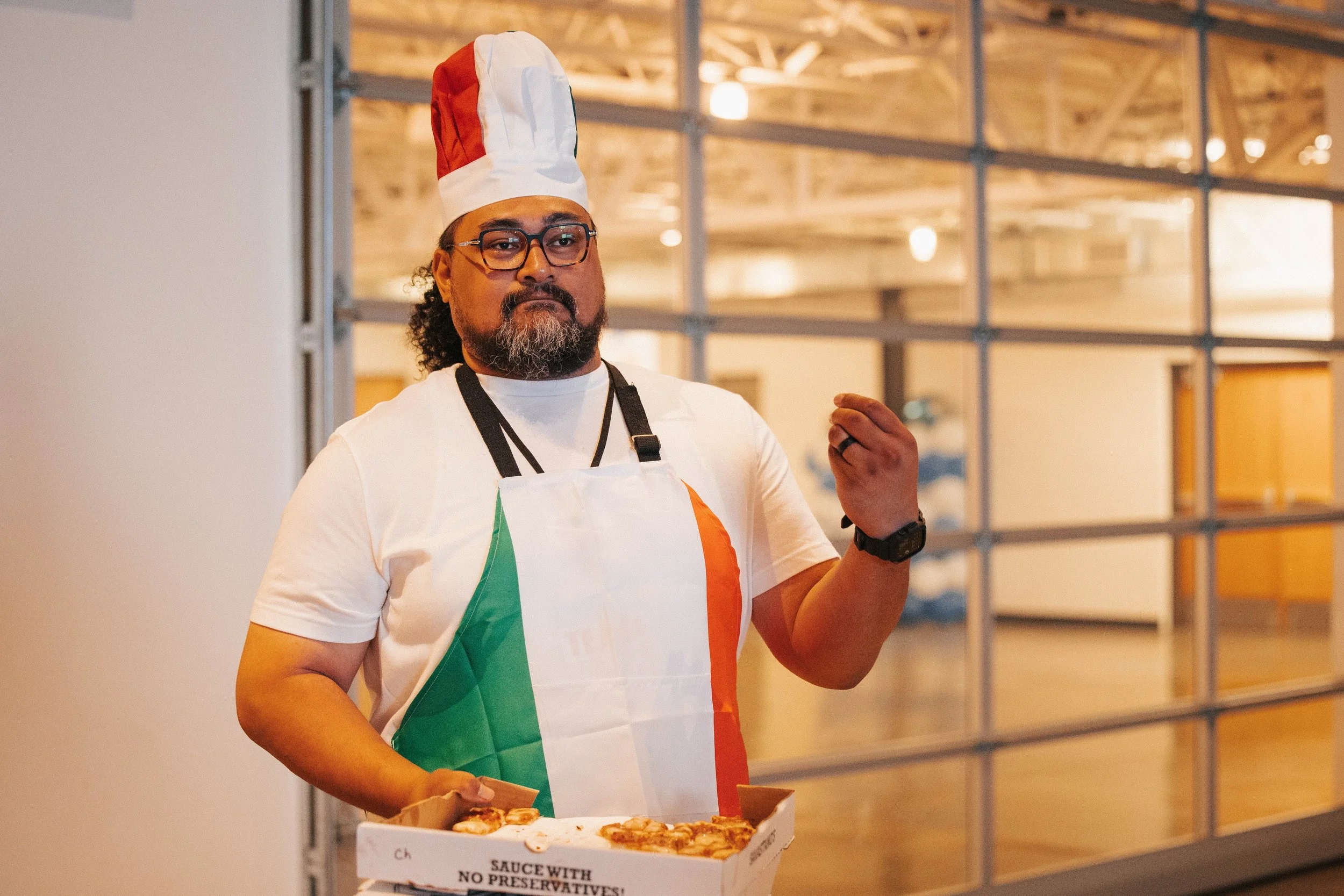 A man wearing a chef's hat and apron with the Italian flag colors holding a pizza in a pizza box, standing in an indoor setting.