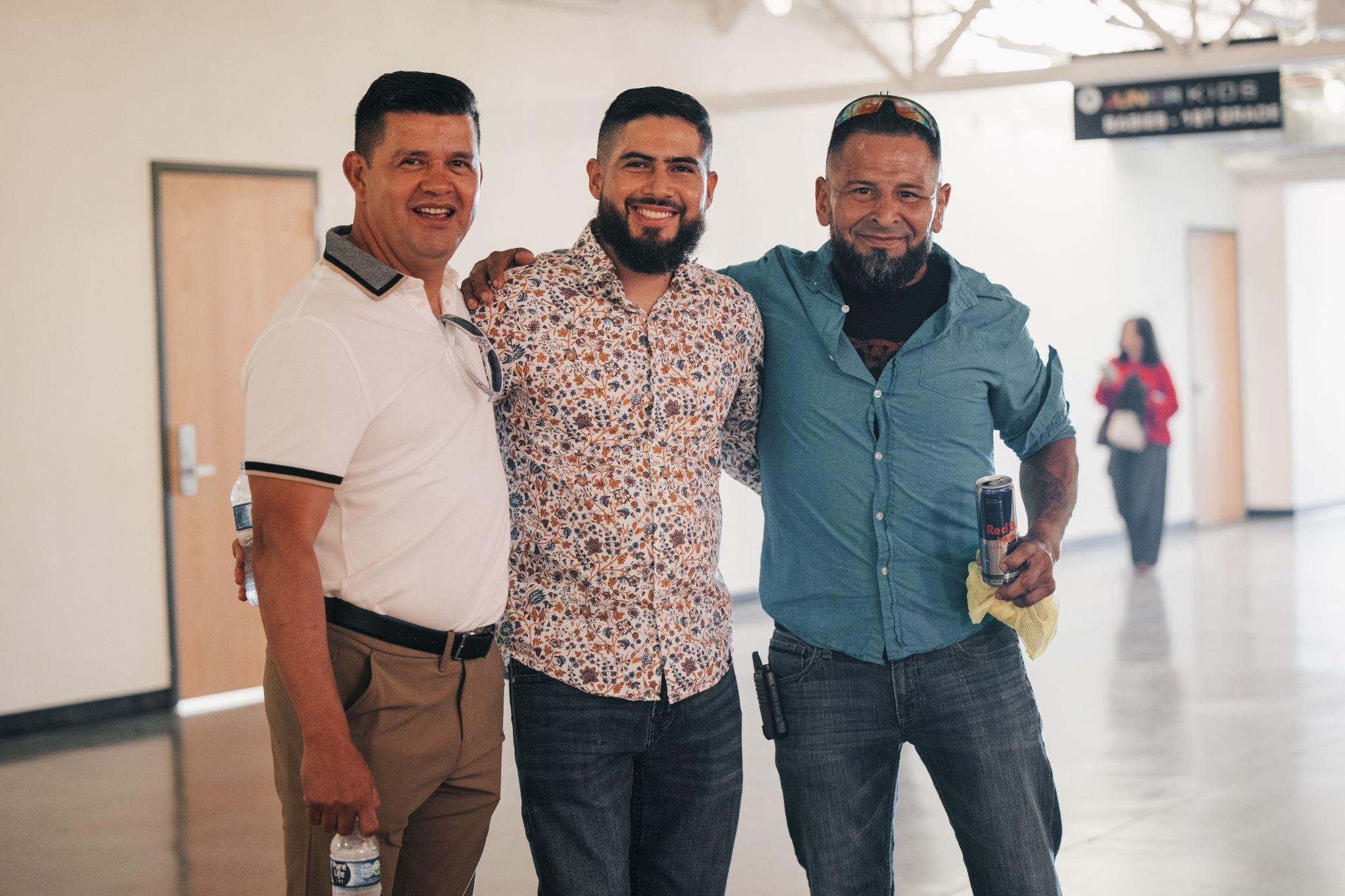 Three men standing together in an indoor hallway, smiling at the camera. The man on the left is holding a water bottle, the man on the right has a can of Red Bull and a yellow cloth in his hand. The man in the middle is wearing a floral shirt, and the others are dressed casually.