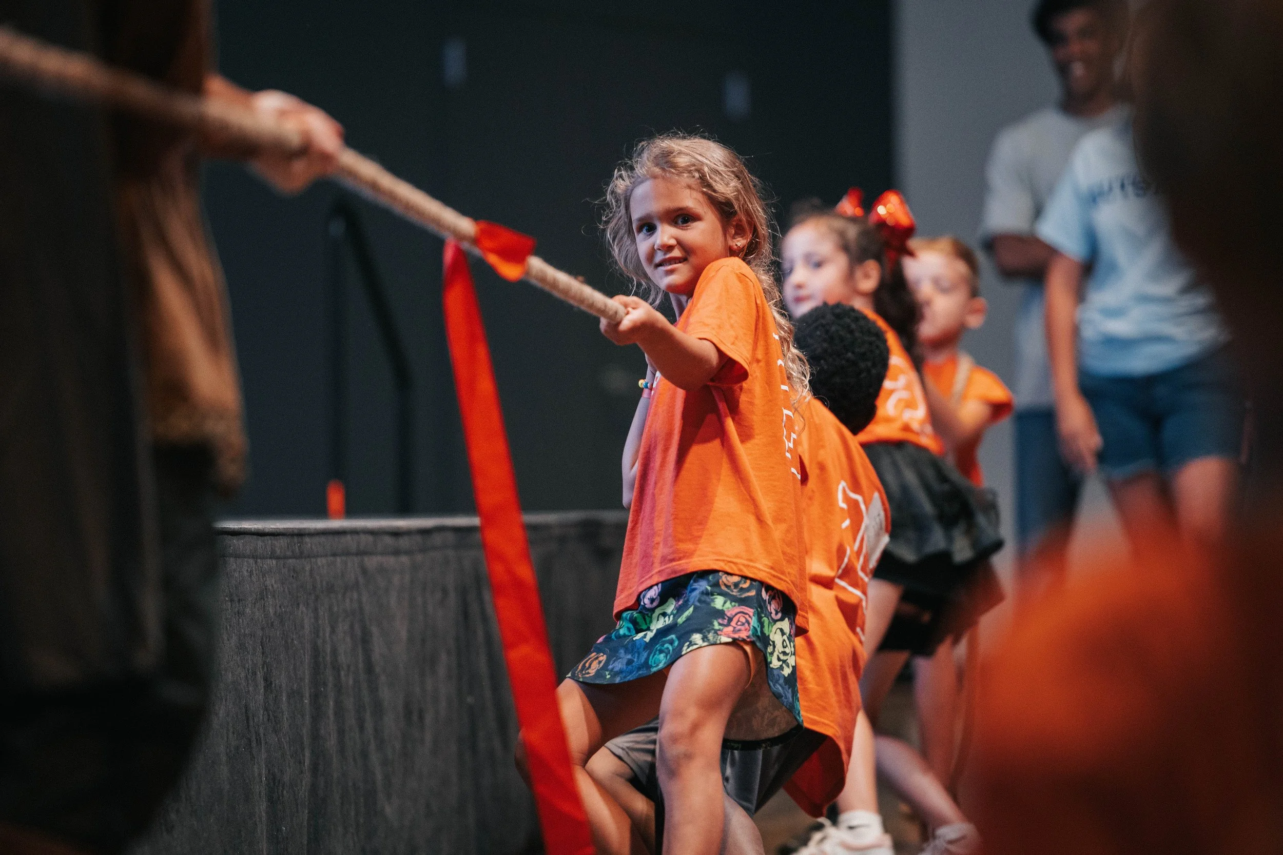 Children in orange shirts pulling on a tug-of-war rope with an adult supervising in the background.