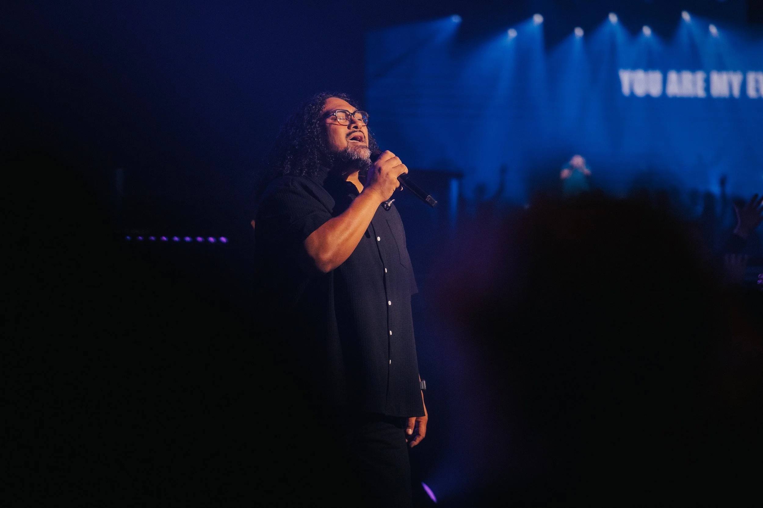 A woman singing on stage with her arm raised, accompanied by musicians, in a church or concert setting. Behind her, a large screen displays the message, "THERE'S NO ONE LIKE OUR GOD," with stage lighting creating a vibrant atmosphere.