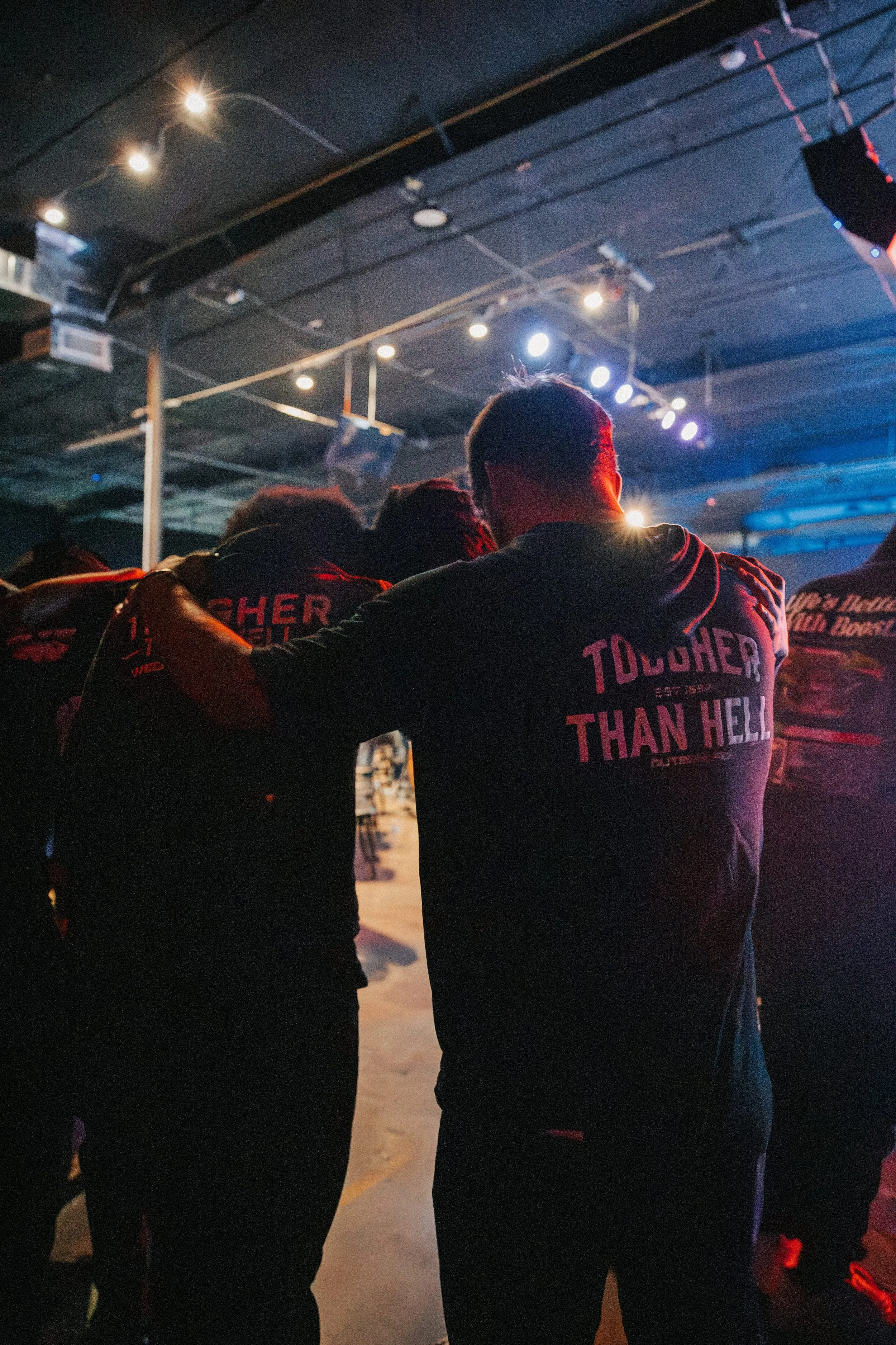 Several people are standing underneath a low-lit ceiling with visible pipes and spotlights, some wearing hoodies with text, in what appears to be an indoor venue or event space.