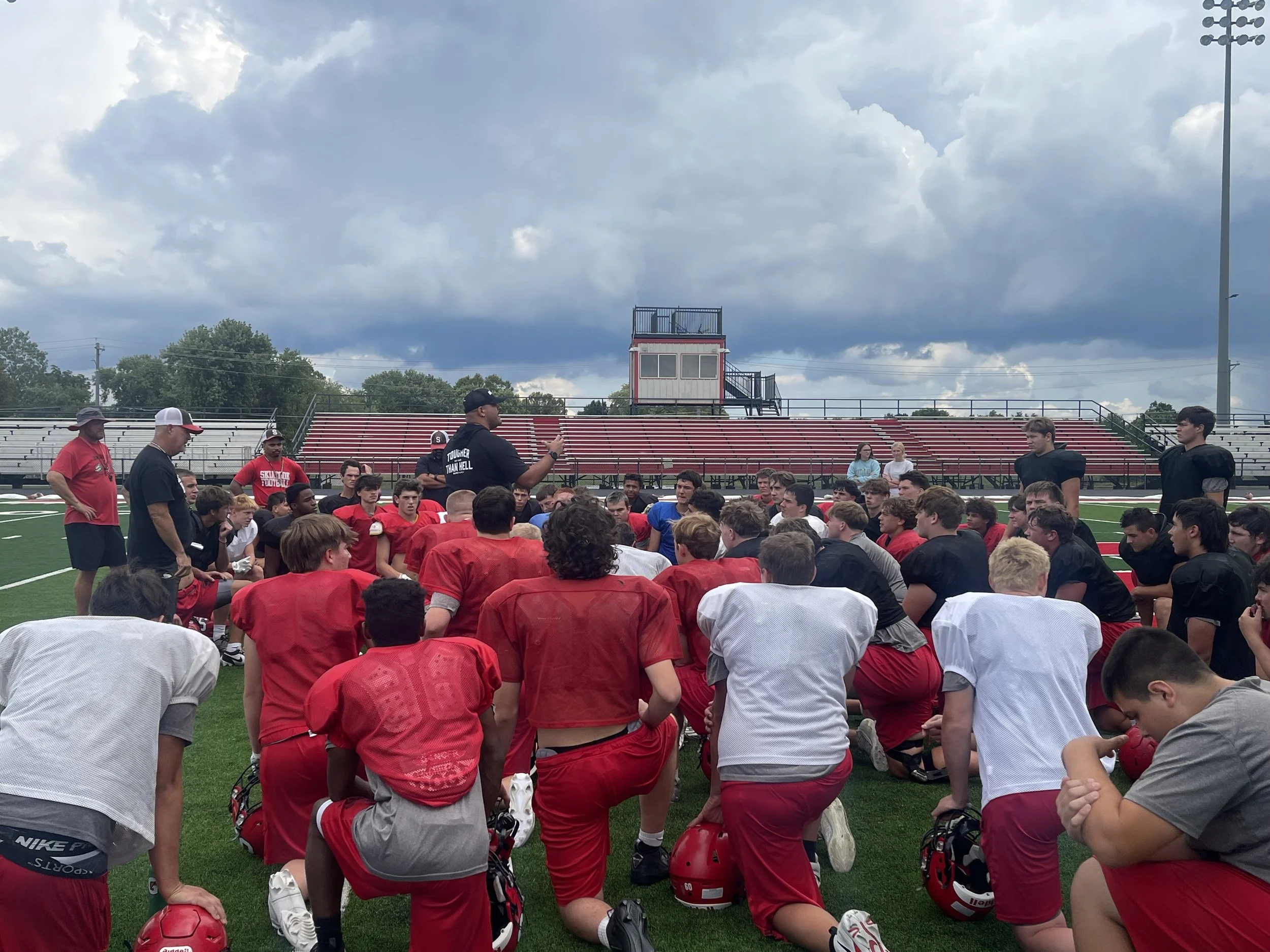 Football players kneeling on field during pre-game or team meeting, with coaches and spectators in background under cloudy sky.