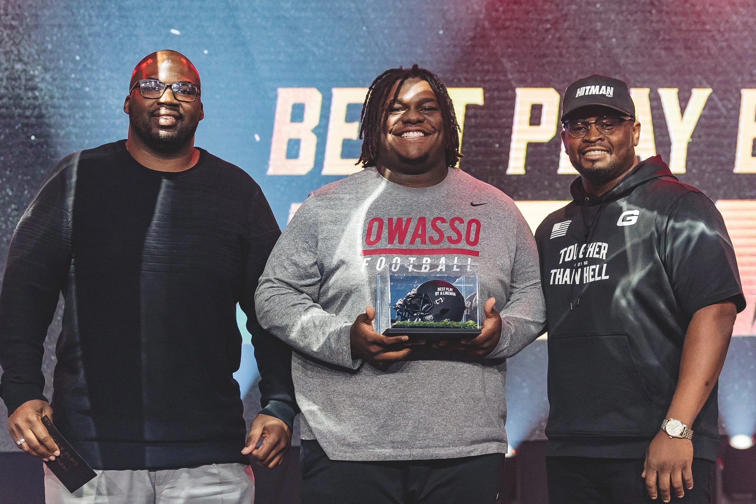 Three men standing together on stage, one holding a football helmet encased in a glass display with a plaque, with a screen in the background displaying the words 'BEAT POWER'.