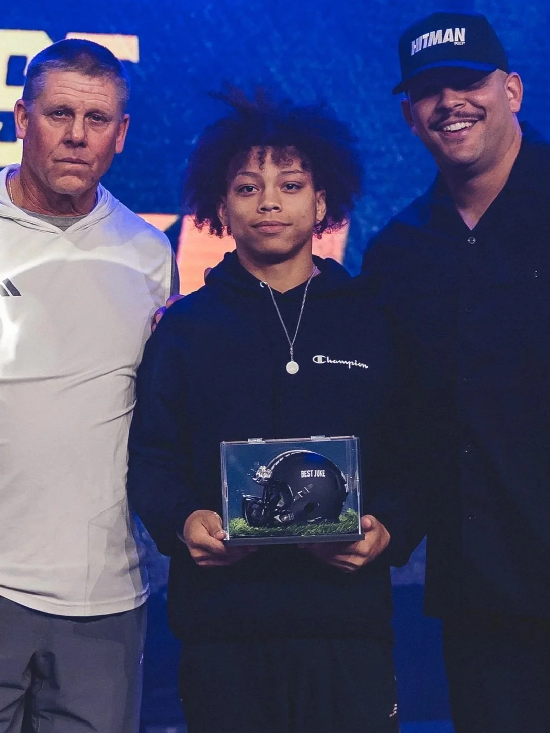 Young boy holding a transparent display case with a black football helmet labeled 'Best Juke,' flanked by two men on a stage.