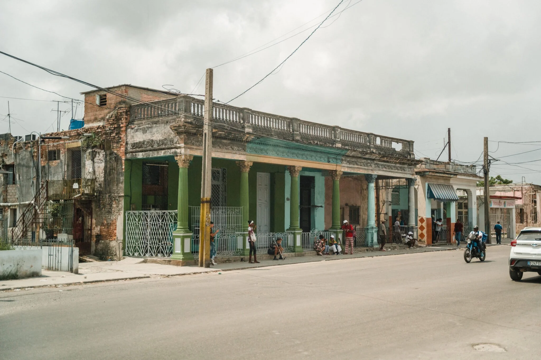 Street scene with old, colorful building with columns and people sitting and standing outside, overcast sky.