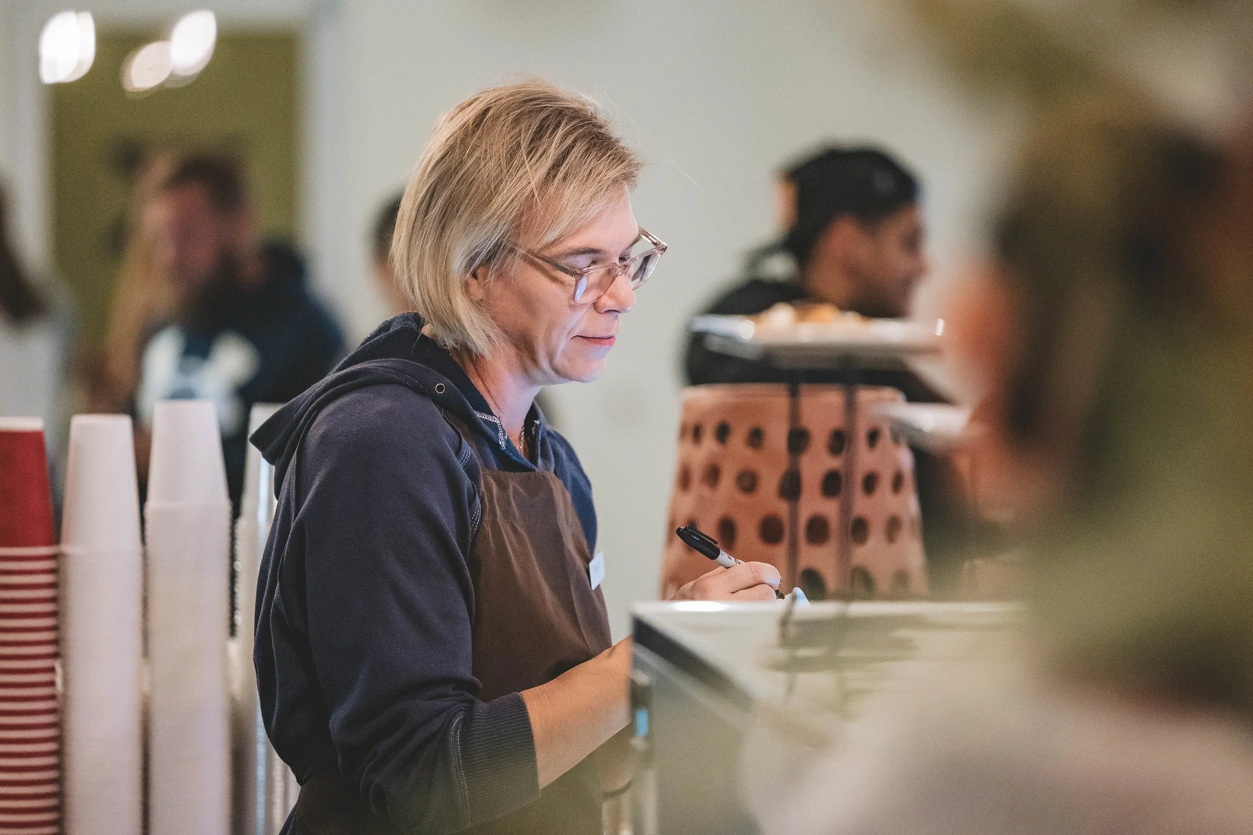 A woman with glasses and blonde hair, wearing a dark hoodie and a brown apron, is writing with a black marker at a counter in what appears to be a cafe or food service setting.