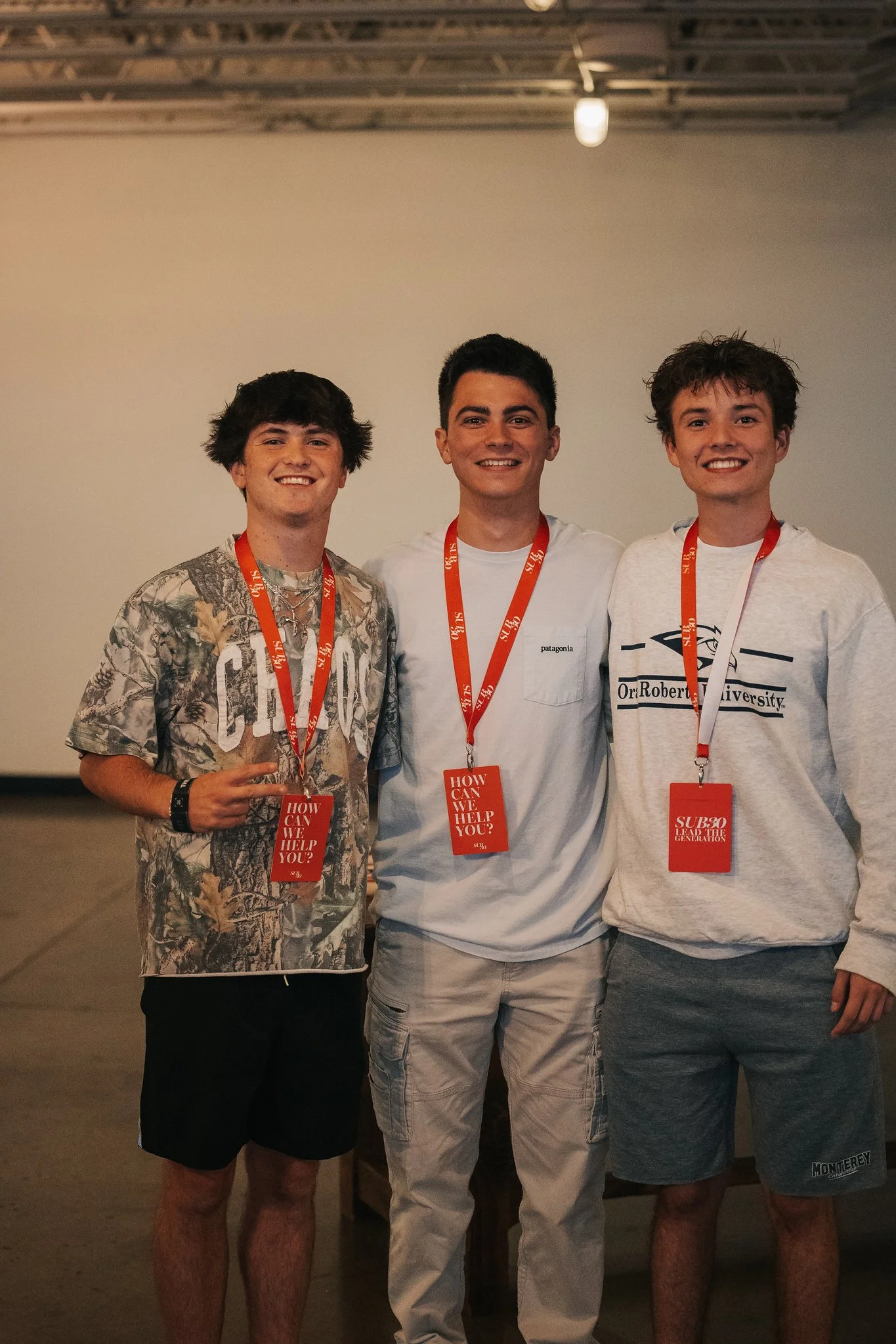 Three young men standing together indoors, each wearing an orange lanyard with a red name tag and smiling at the camera.