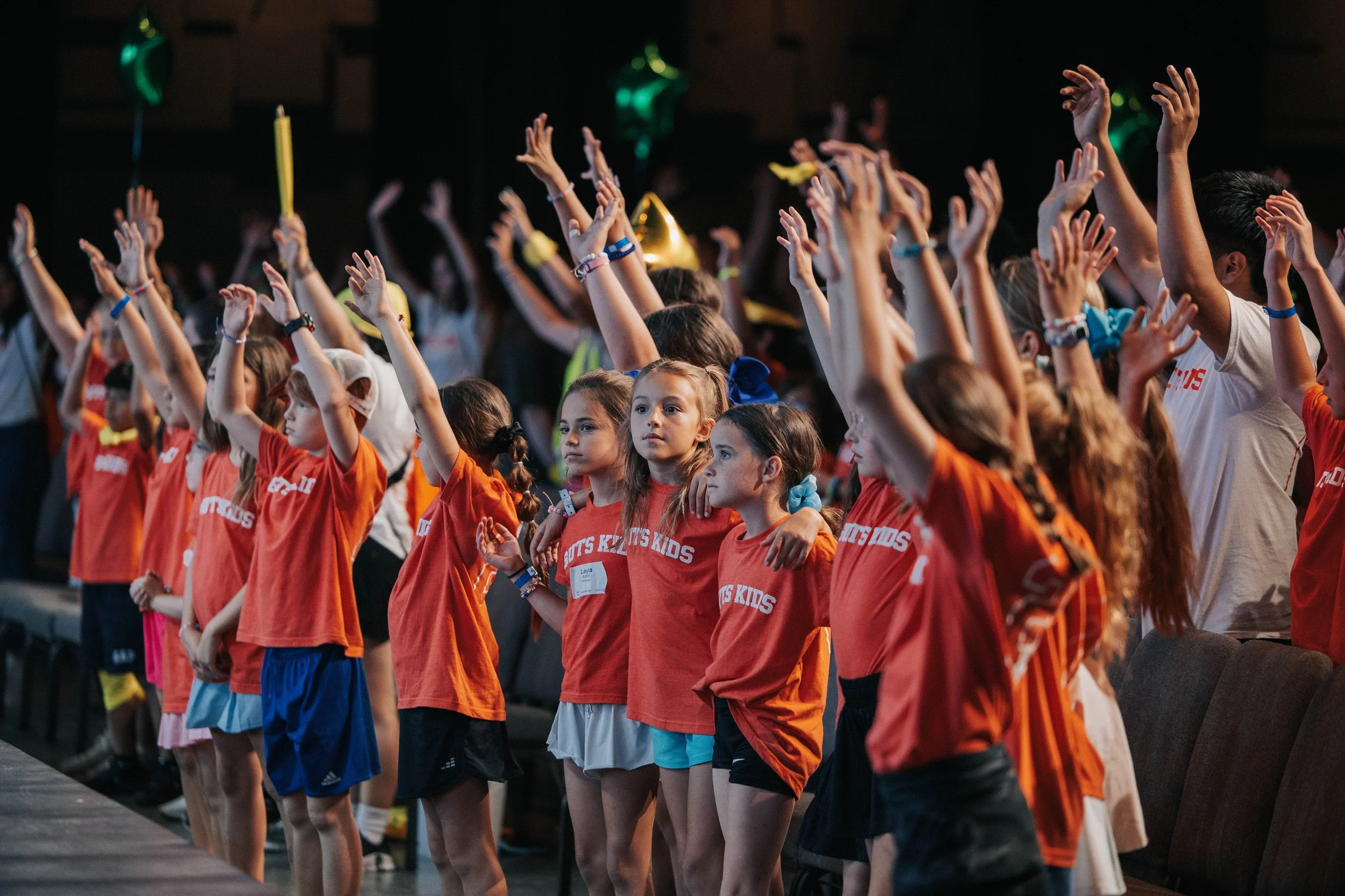 Group of young children wearing orange shirts, standing in a row with arms raised, participating in an indoor event.
