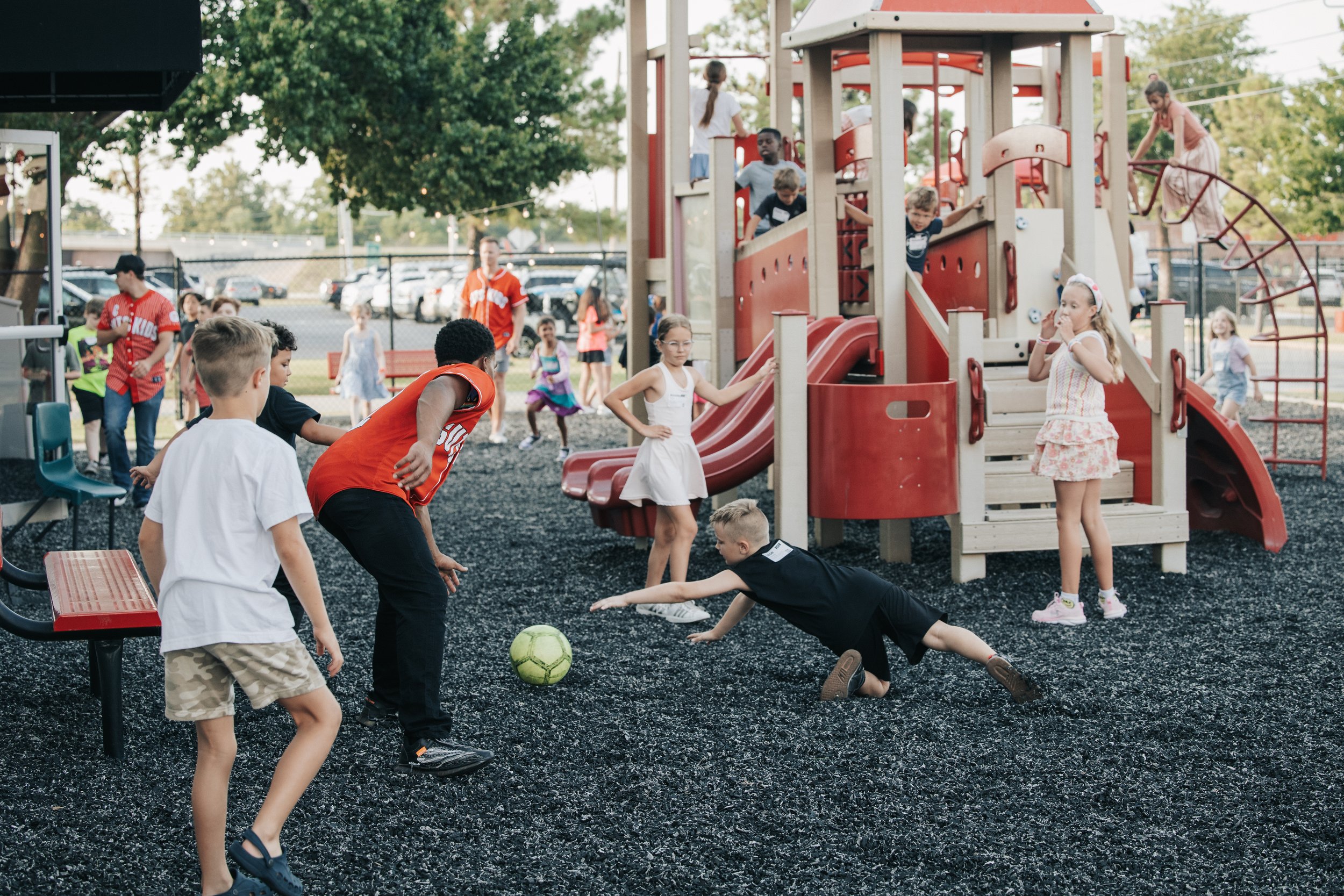 Children playing soccer on a playground with a red and beige play structure, while other kids and adults are in the background.