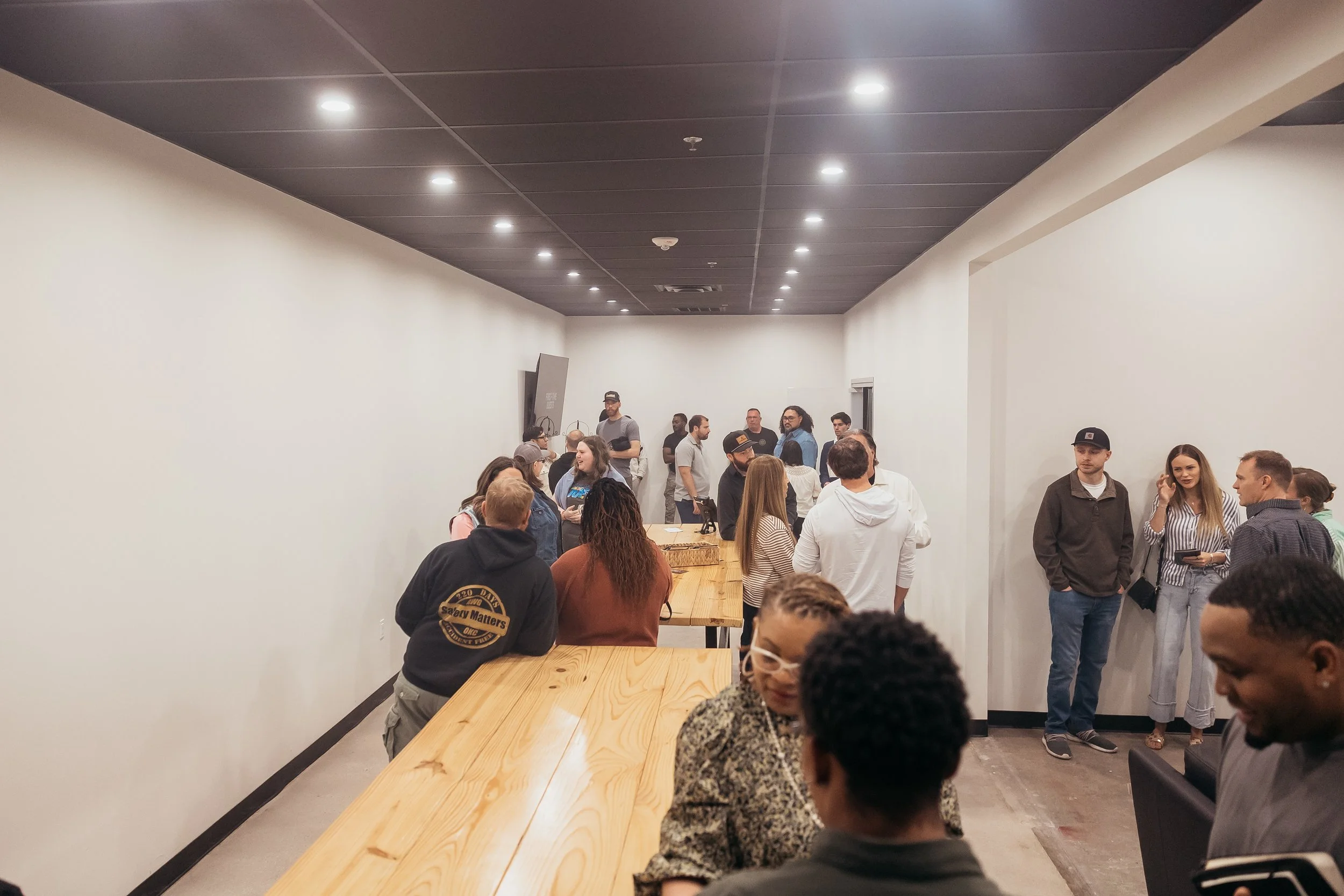 People socializing in a room with white walls, some standing and others sitting at a large wooden table, with a dark ceiling and bright ceiling lights.