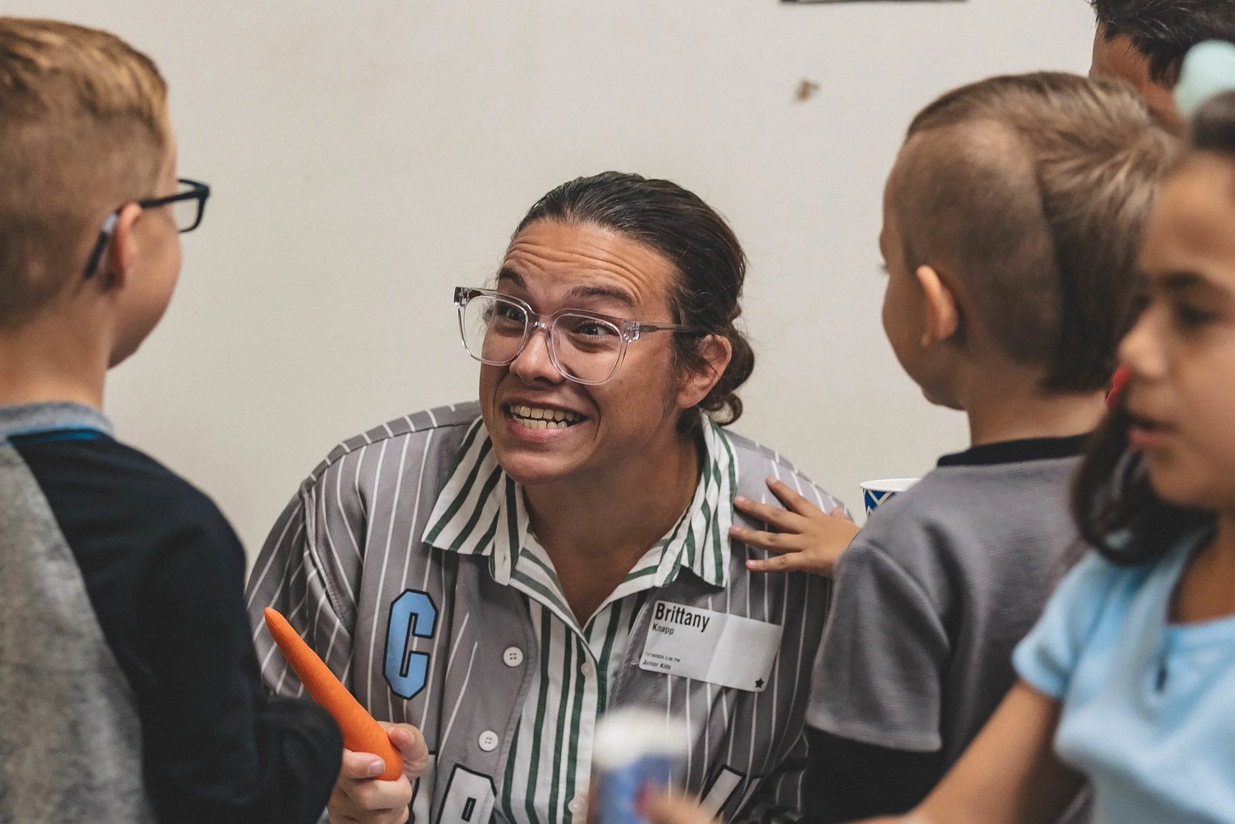 A woman with glasses, wearing a striped shirt and hispanic name tag, is surrounded by children, some holding cups and one with a carrot, having an animated conversation indoors.