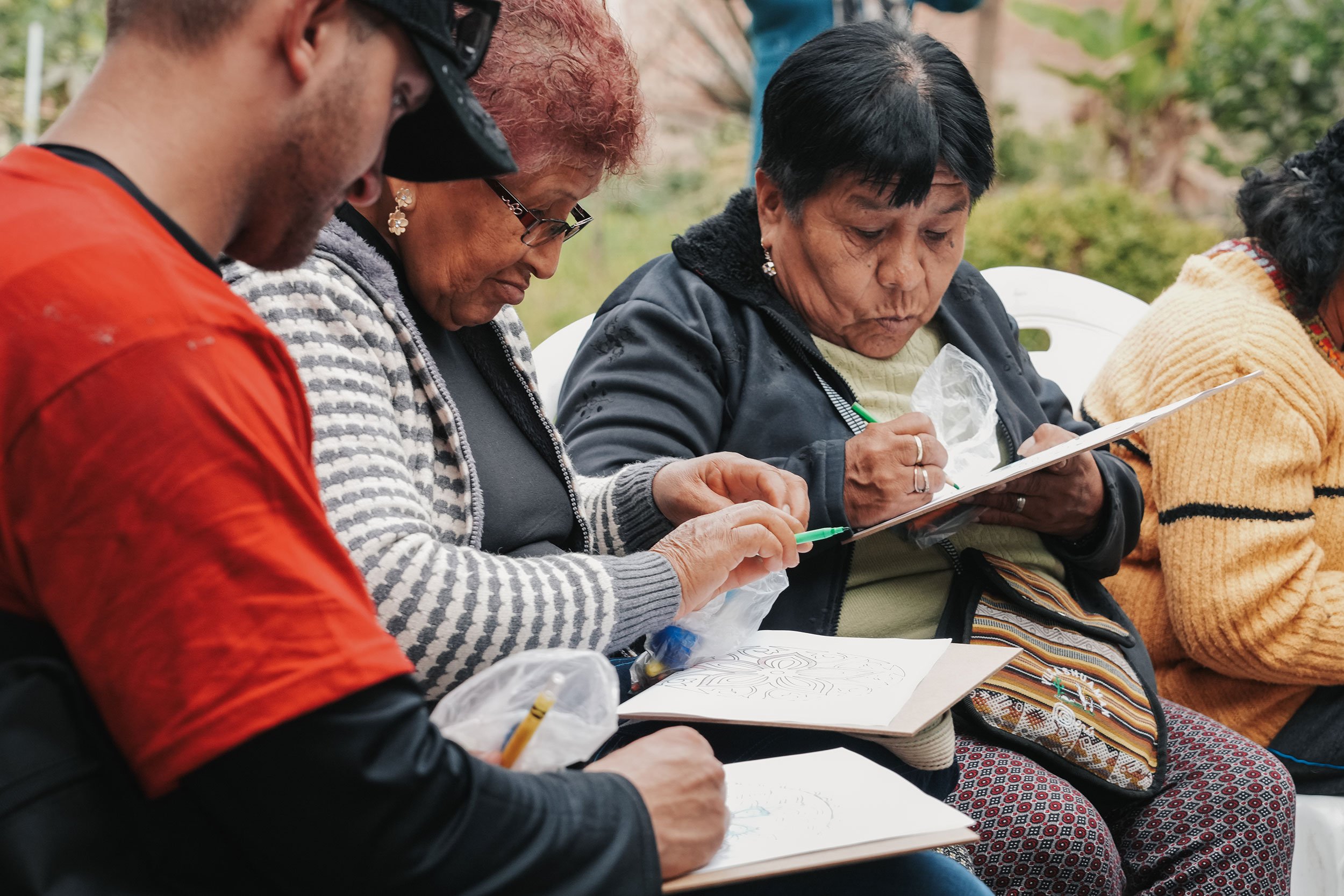 Group of older women and a young man sitting outdoors, focused on drawing and writing in notebooks.
