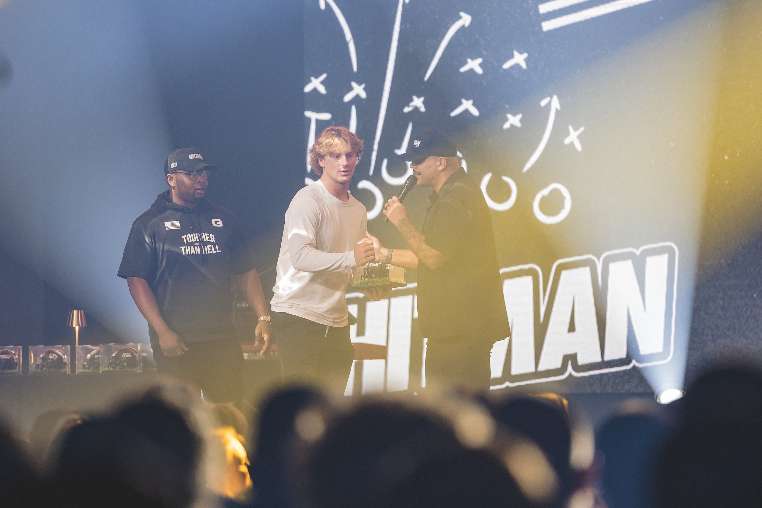 Three men standing on a stage during an award ceremony, with a large screen behind them displaying text and graphics. One man is holding a microphone and an award, while the others watch. The audience is visible in the foreground.
