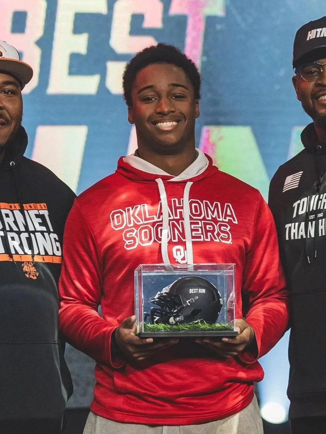 A young man in a red Oklahoma Sooners hoodie is holding a display case with a black football helmet labeled 'Best Run.' He is smiling and standing between two men, one in a black hoodie and the other in a black jacket and cap. The background features colorful text that says 'BEST' and 'WIN.'