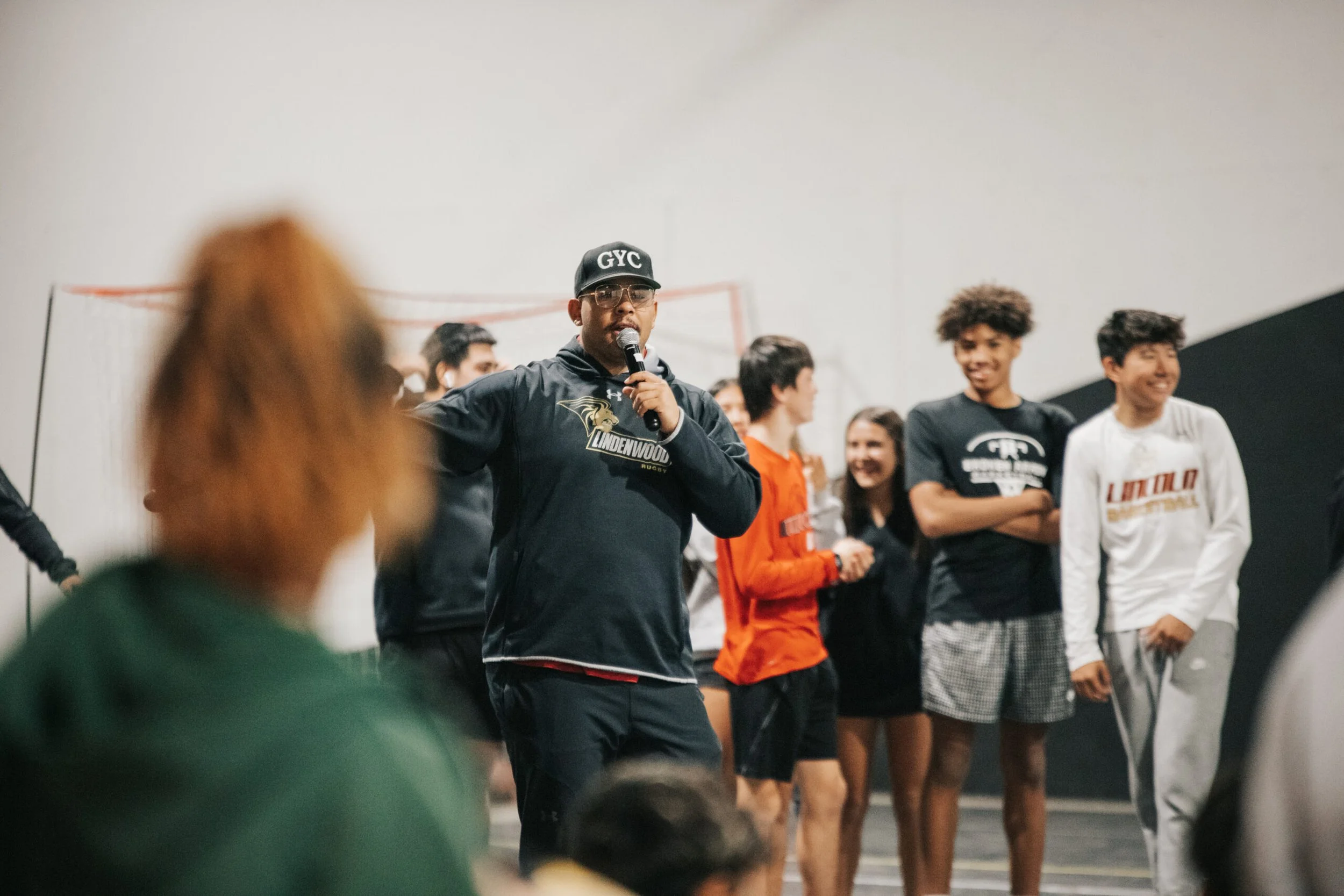 A man wearing glasses, a cap with 'GYC' on it, and a black jacket, is speaking into a microphone in front of a group of young people. The group appears to be in a gymnasium or indoor sports facility, with some people smiling and engaging with the speaker.