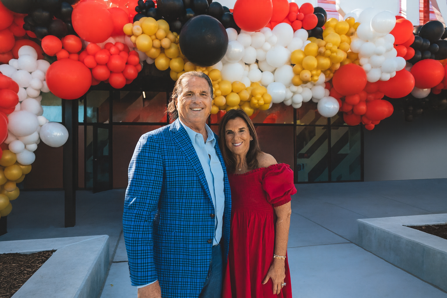 A smiling man and woman standing together outdoors in front of a building decorated with colorful balloons. The man is wearing a blue checked blazer over a white shirt, and the woman is dressed in a red off-the-shoulder dress.