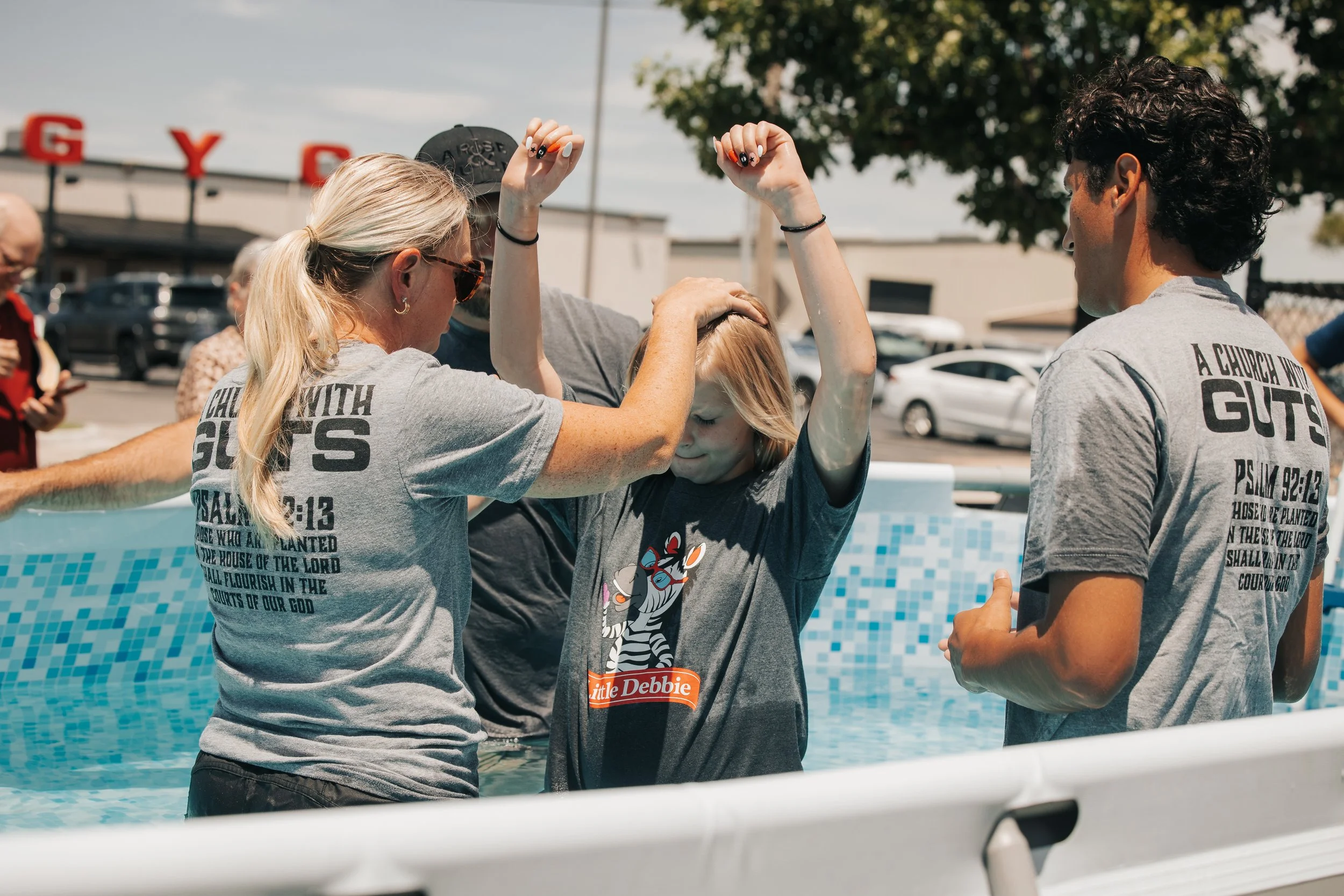 A baptism ceremony taking place outdoors in a swimming pool, with a woman and a young girl being baptized, surrounded by other people and cars in the background.