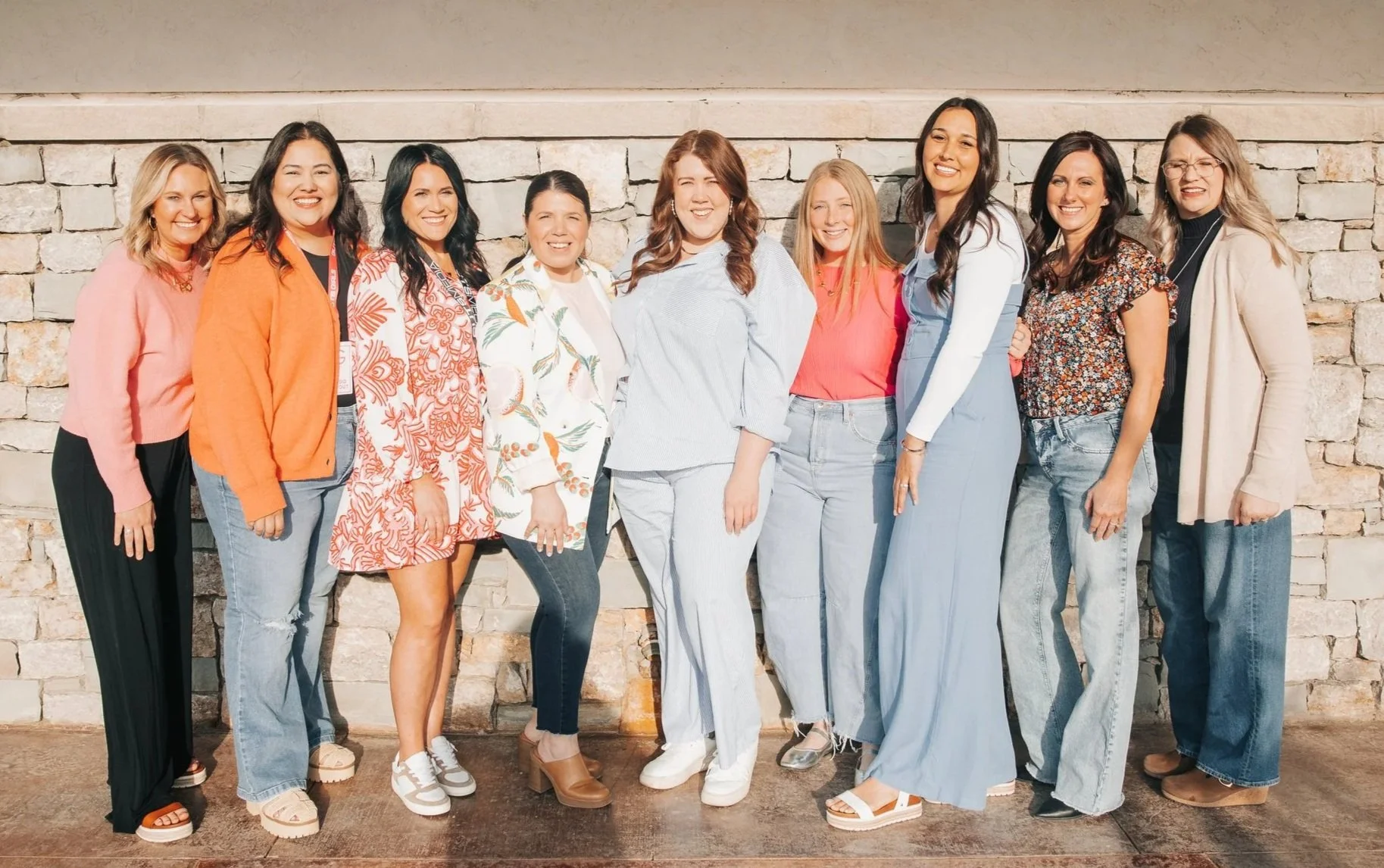 Group of eleven women standing outdoors against a stone wall, smiling.