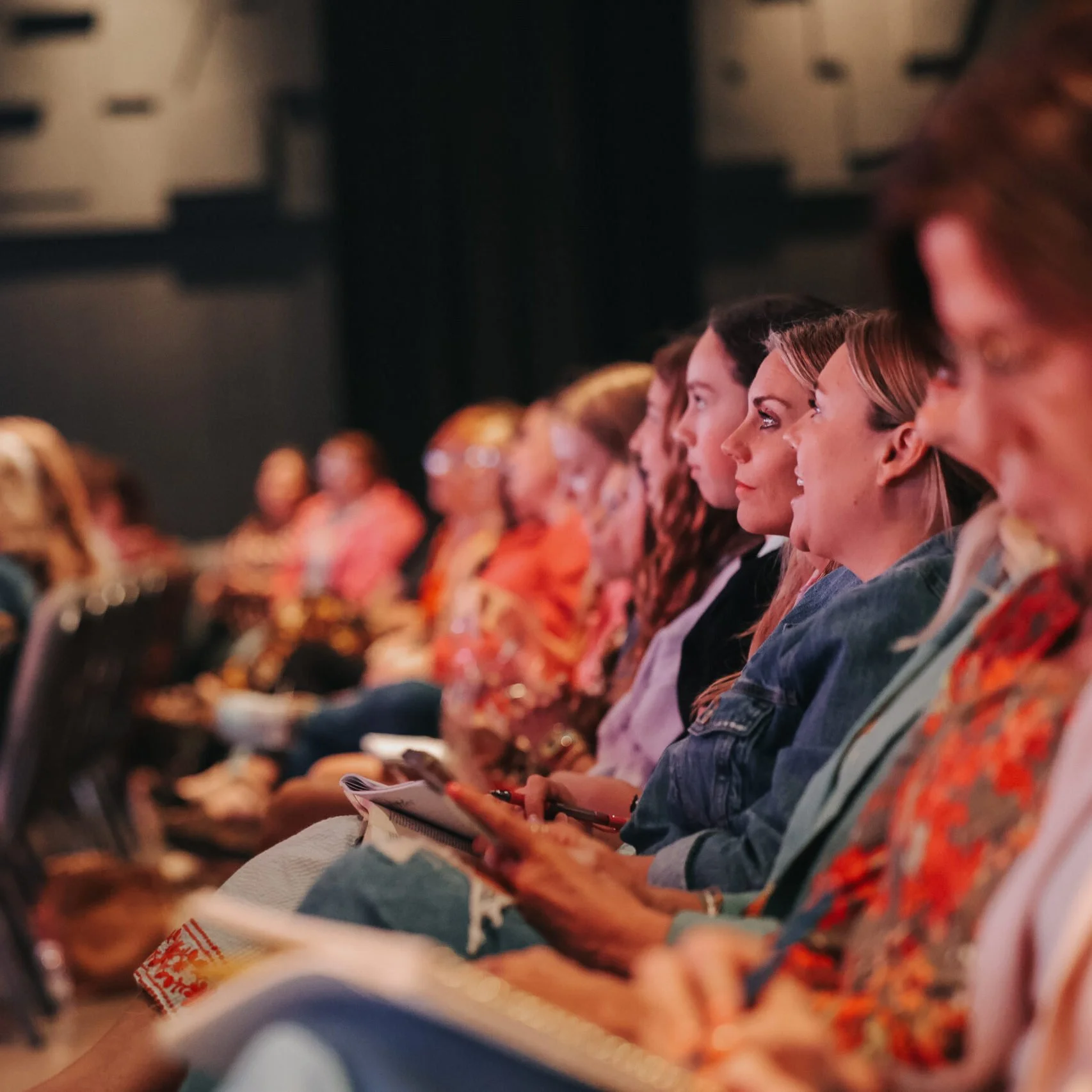 Audience of women sitting in a dark auditorium, attentively listening to a presentation or performance.