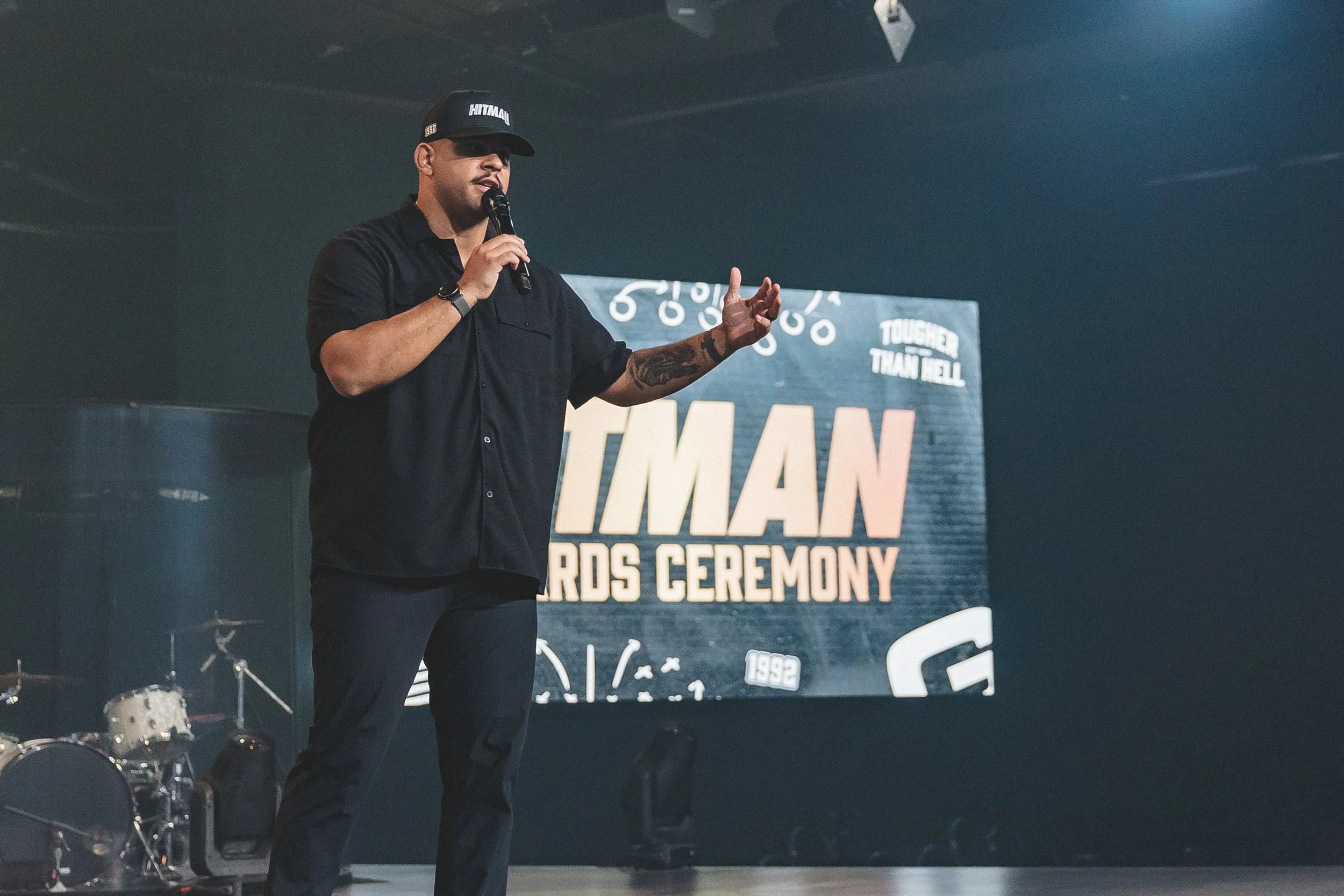 A man wearing a black cap, black shirt, and black pants speaking into a microphone on stage during an awards ceremony, with a large sign behind him that reads 'Tougher Than Hell.'