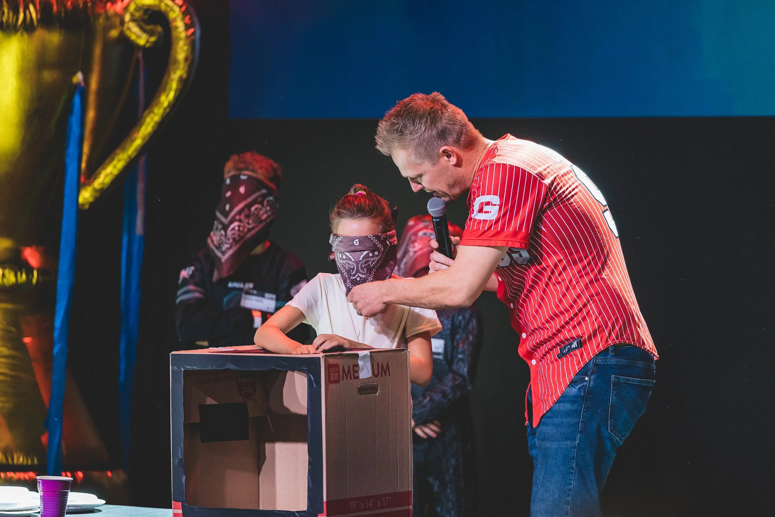 A man showing a girl how to use a microphone during an event, with people in masks and dark background.
