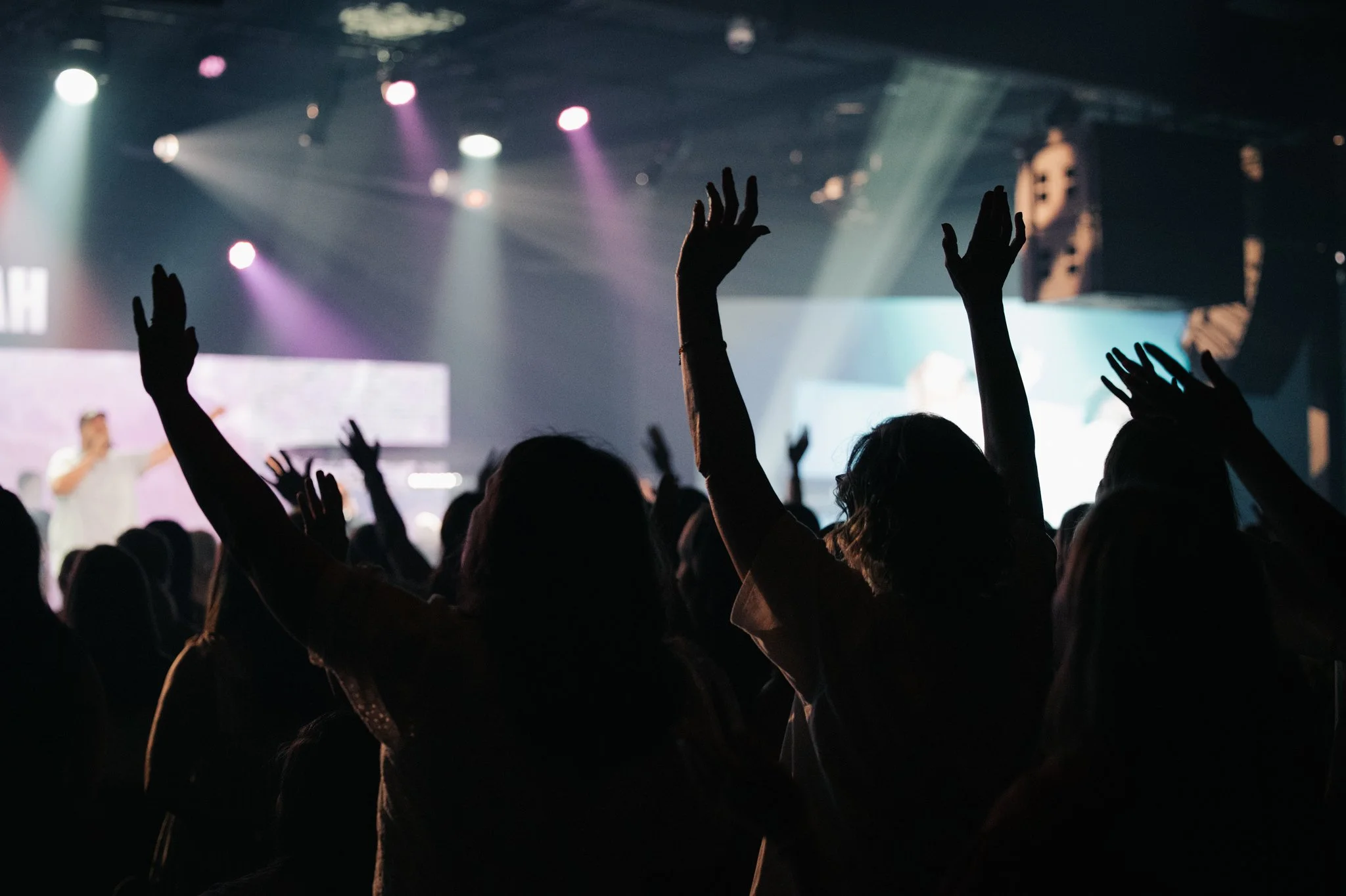 Crowd of people raising their hands at a concert or event with stage lights and a performer in the background.