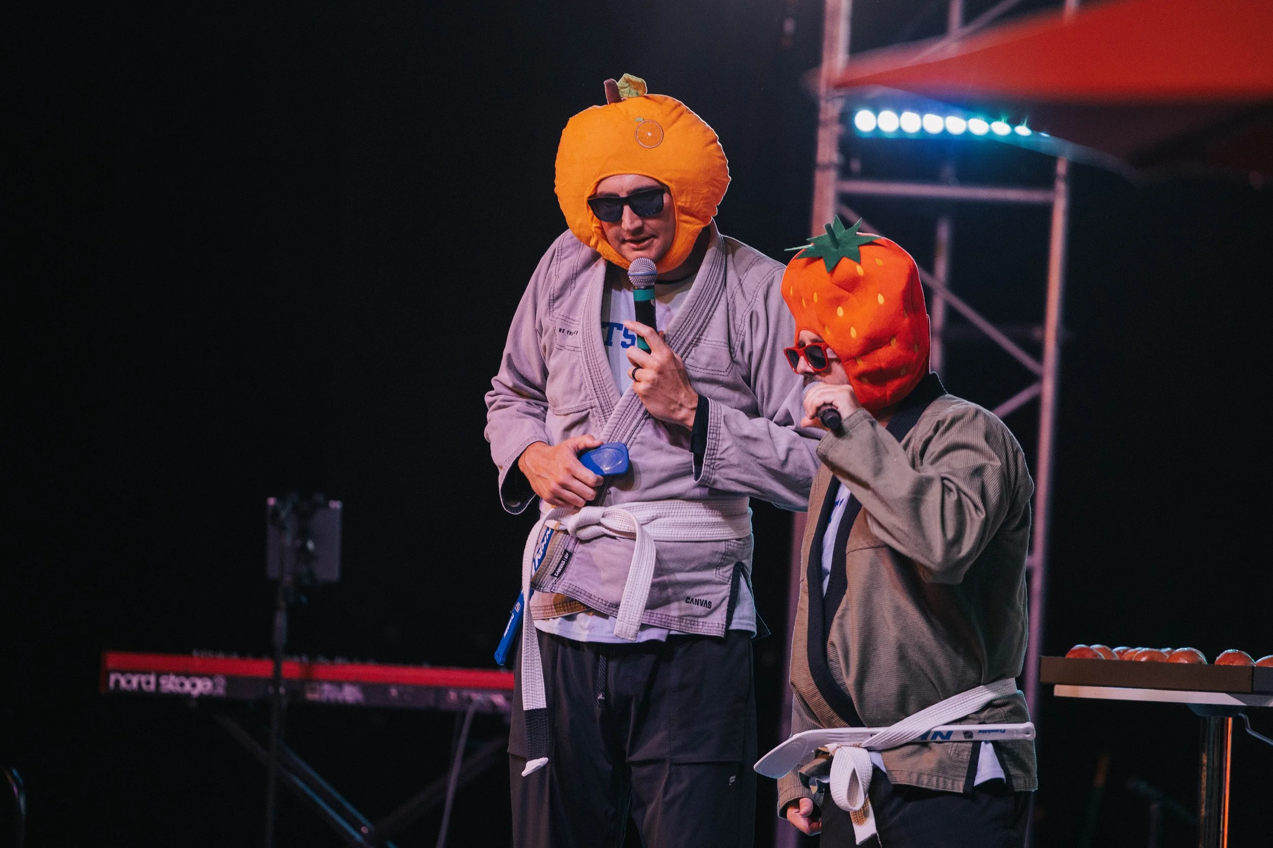Two men wearing Halloween pumpkin head costumes and sunglasses are on stage, holding microphones and speaking to each other, with musical equipment in the background.