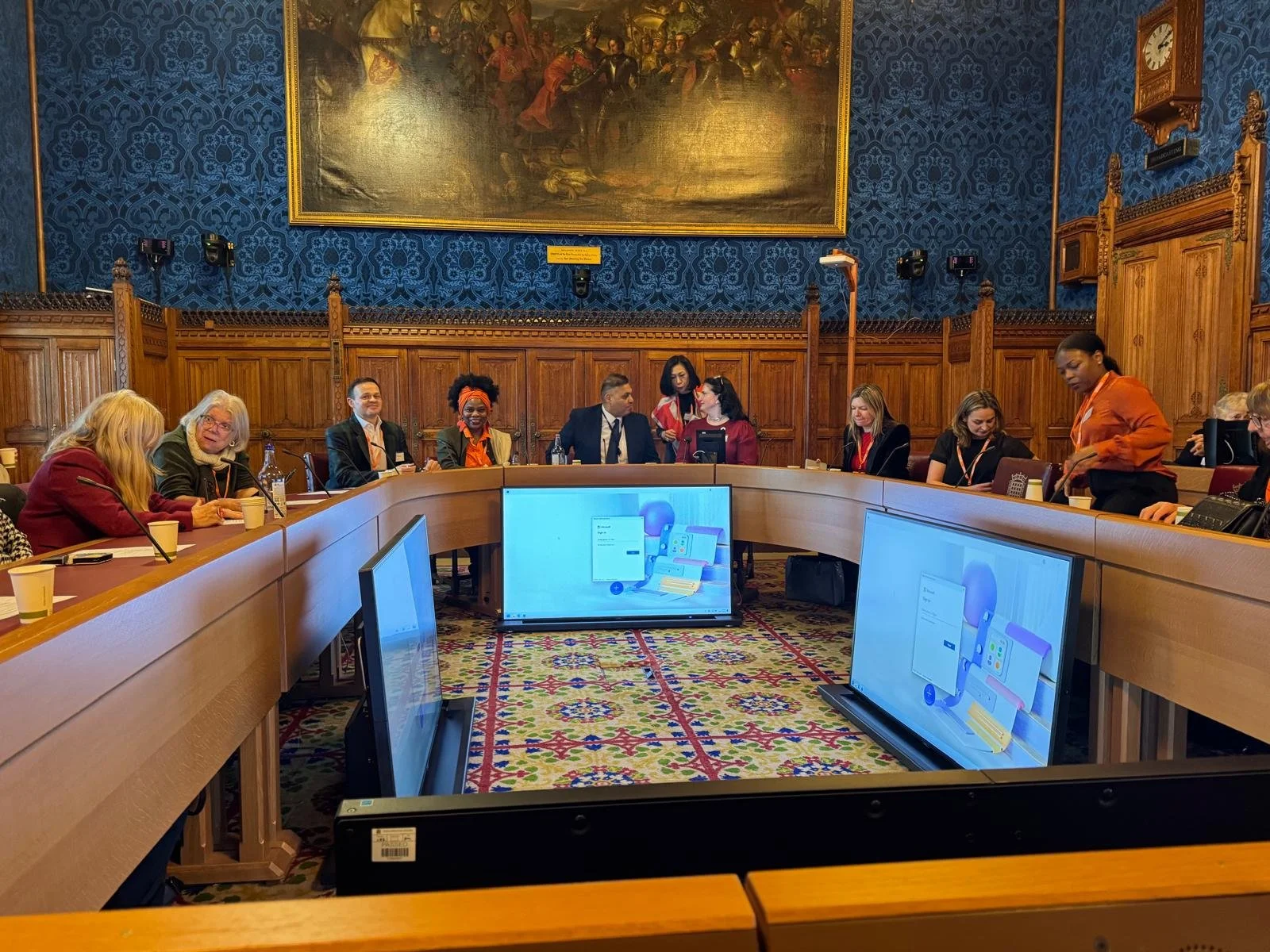 A group of diverse people sitting around a U-shaped conference table in a large, ornate room with wooden paneling, a large painting on the wall, and several monitors on the floor displaying digital graphics.