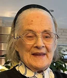 An elderly woman wearing glasses, a black headband, and a striped scarf, smiling gently indoors.