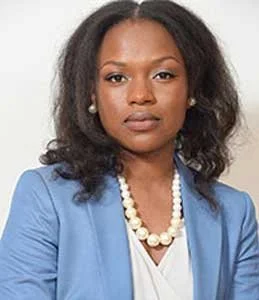 A woman with dark curly hair wearing a blue blazer, white blouse, a pearl necklace, and pearl earrings, looking at the camera with a serious expression.