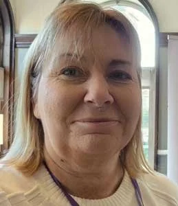 A woman with blonde hair smiling inside a train or bus, with windows and seats visible in the background.
