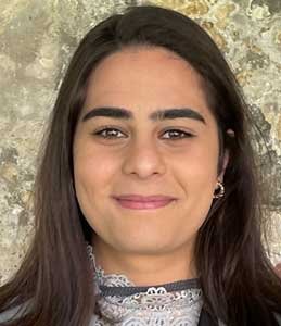 Close-up portrait of a young woman with long dark hair, dark eyebrows, and a slight smile, standing against a textured beige wall.