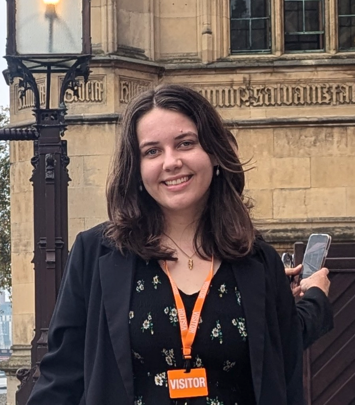 A smiling young woman with shoulder-length dark hair, wearing a black floral dress, a black blazer, and an orange visitor badge around her neck. She is standing outdoors in front of a historic stone building with stained glass windows and ornate architecture, holding a smartphone in one hand.