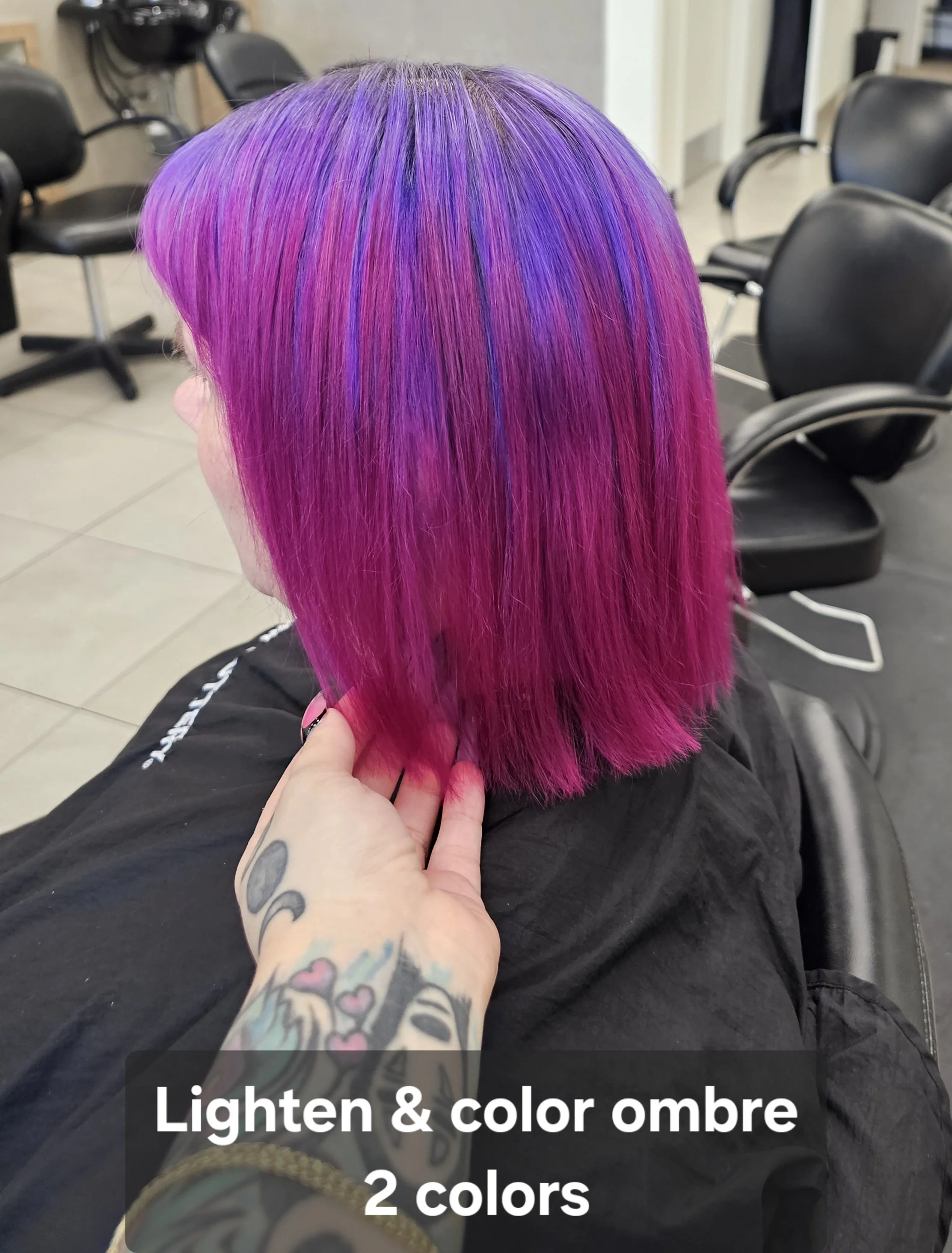 Side view of a person with freshly styled, vibrant pink and purple ombre hair, seated in a salon chair, with a tattooed hand touching the hair and a caption indicating lighten and color ombre with two colors.