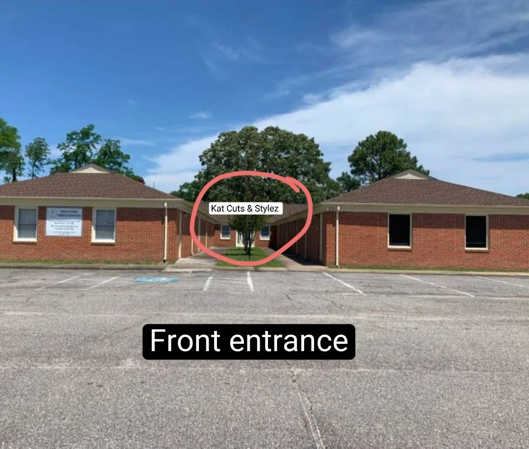 A brick building with a parking lot in front, labeled as 'Front entrance.' The building has four windows, and a sign on the left side. There is a pathway leading to a tree in the center, with the text 'Kat Cuts & Stylez' circled in red above it. The sky is partly cloudy.