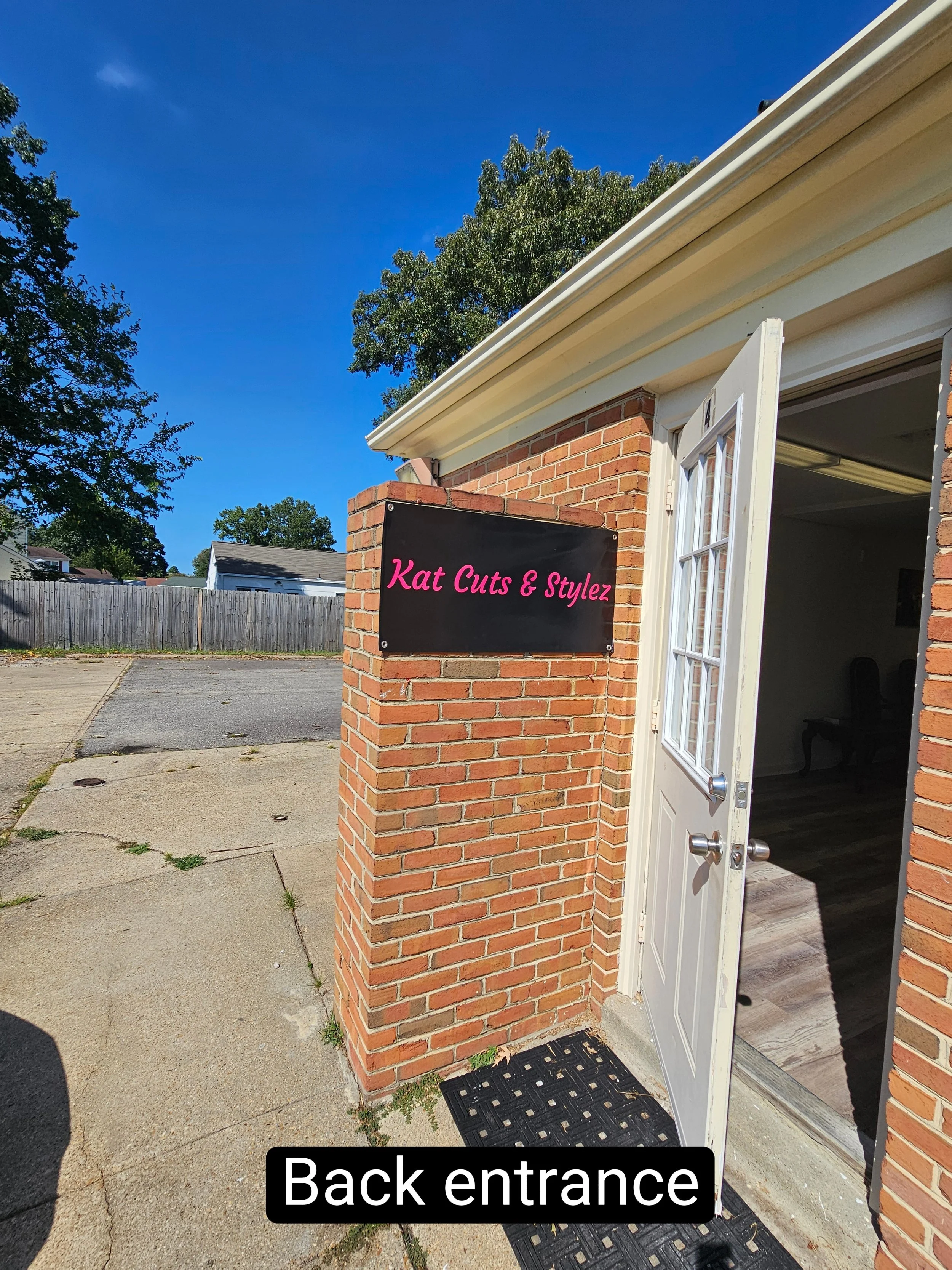 Exterior view of a brick building with a sign that reads "Kat Cuts & Stylez" near the back entrance, which has a white door open, revealing part of the interior.