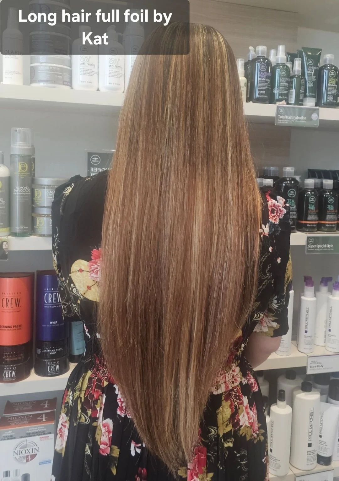 A woman with long, straight, full foil hair stands in front of shelves stocked with hair care products in a store. The hair is light brown with blonde highlights.