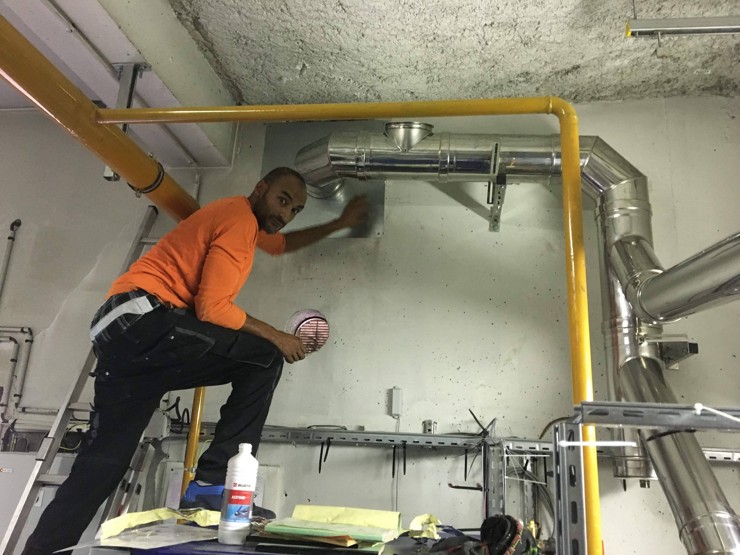 Miel, le technicien en tenue orange inspecte une canalisation d'une ventilation dans une pièce industrielle.
