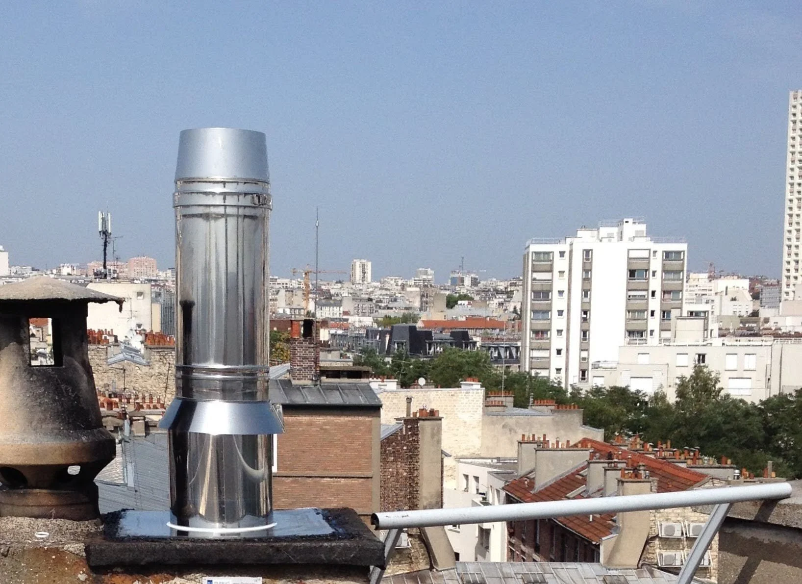 Vue urbaine depuis un toit avec un système d'évacuation en métal, toits en brique, immeubles résidentiels et un ciel clair.