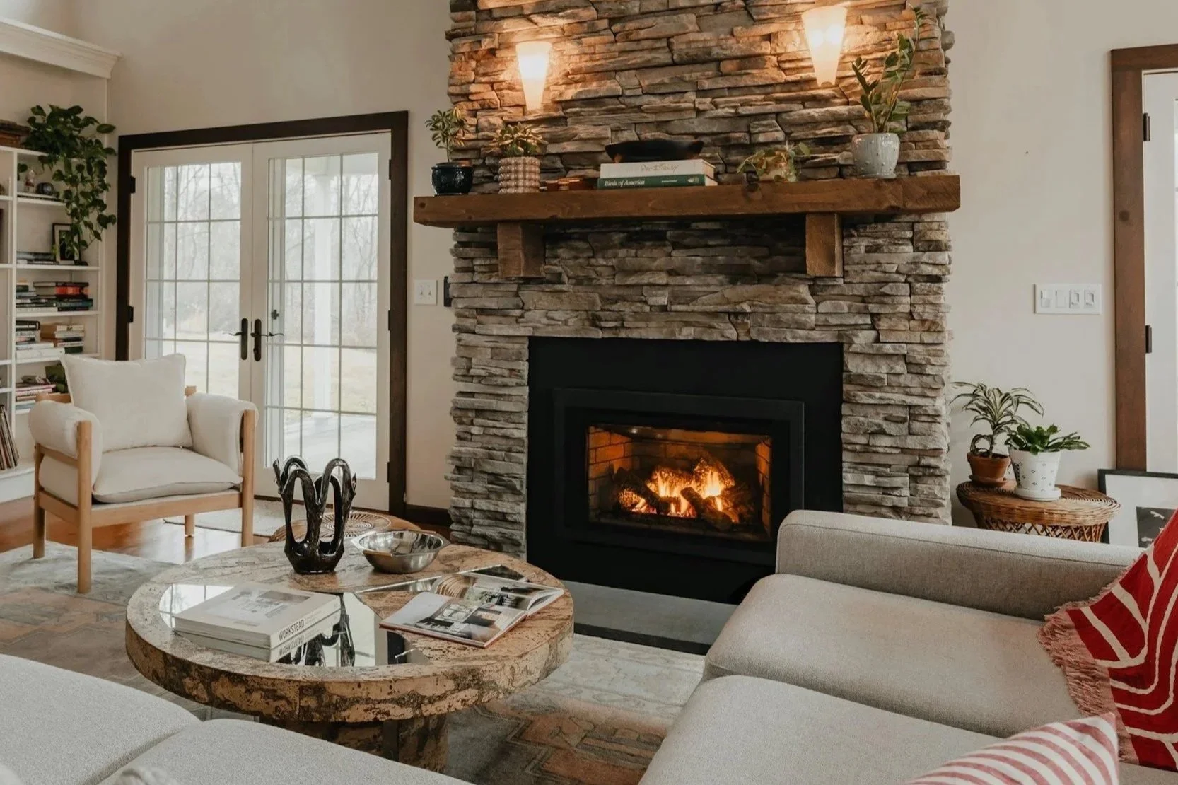 Salle de séjour avec cheminée en pierre, canapé beige, table en bois avec magazines, fauteuil blanc, étagère à livres, plantes décoratives, et porte-fenêtre donnant sur un jardin