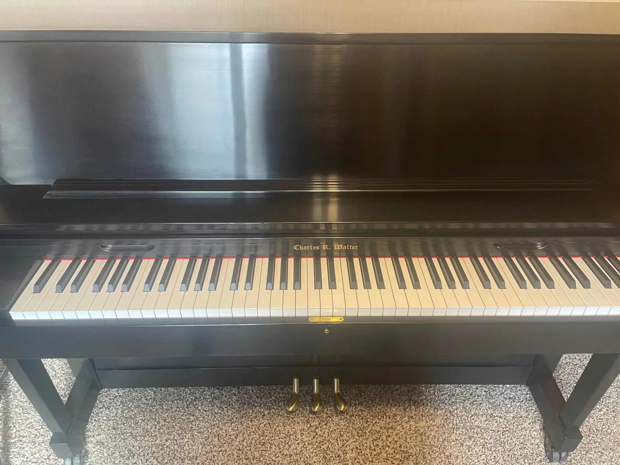 A black upright piano with a gold signature that reads 'Charles R. Walter' on the keyboard cover, resting on a carpeted floor.
