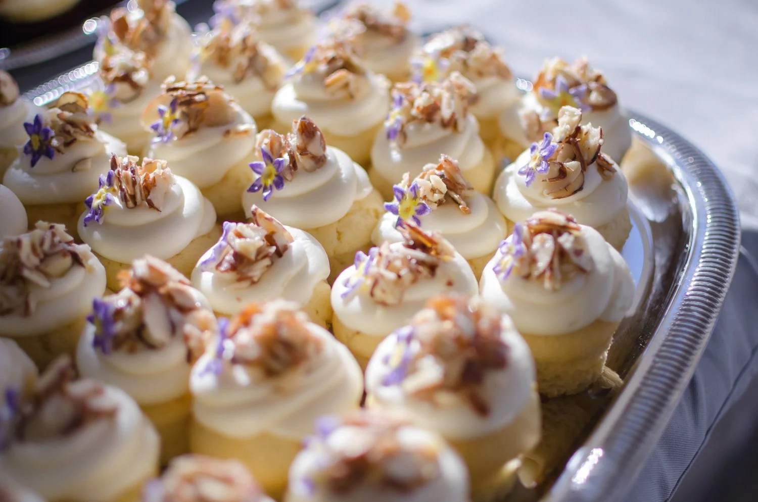 A tray of cupcakes topped with white frosting, chopped nuts, and purple edible flowers.