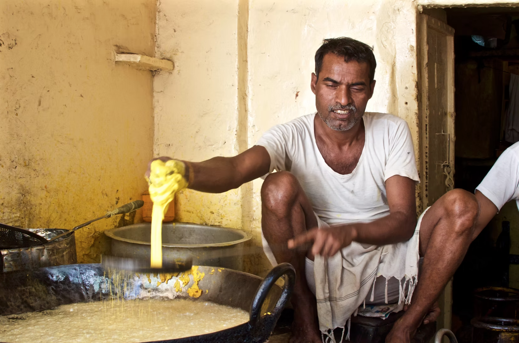 A man cooking food in a traditional kitchen with yellow walls, lifting a piece of cooked food out of a pot.