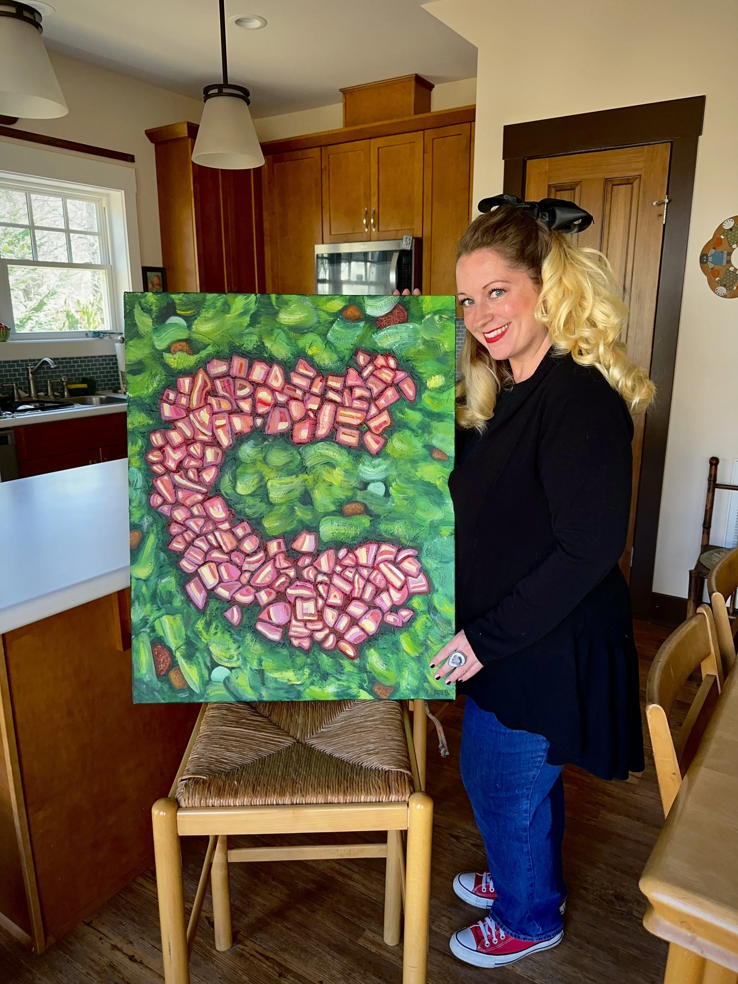 Woman with blonde curled hair, black bow, red lipstick, wearing black cardigan and blue jeans, holding a colorful abstract painting in a kitchen.