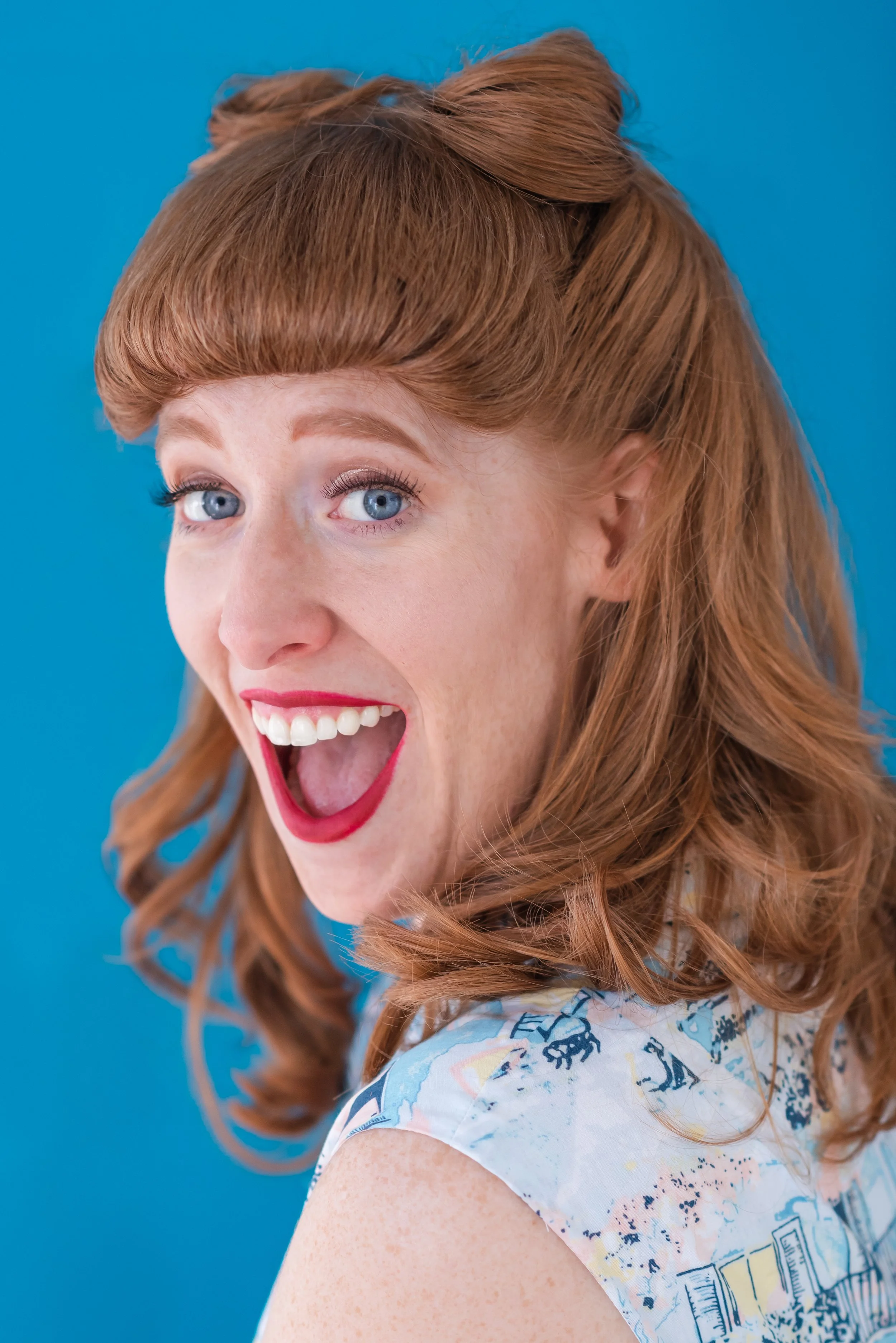 A red-haired woman with blue eyes, smiling widely and showing her teeth, against a blue background.