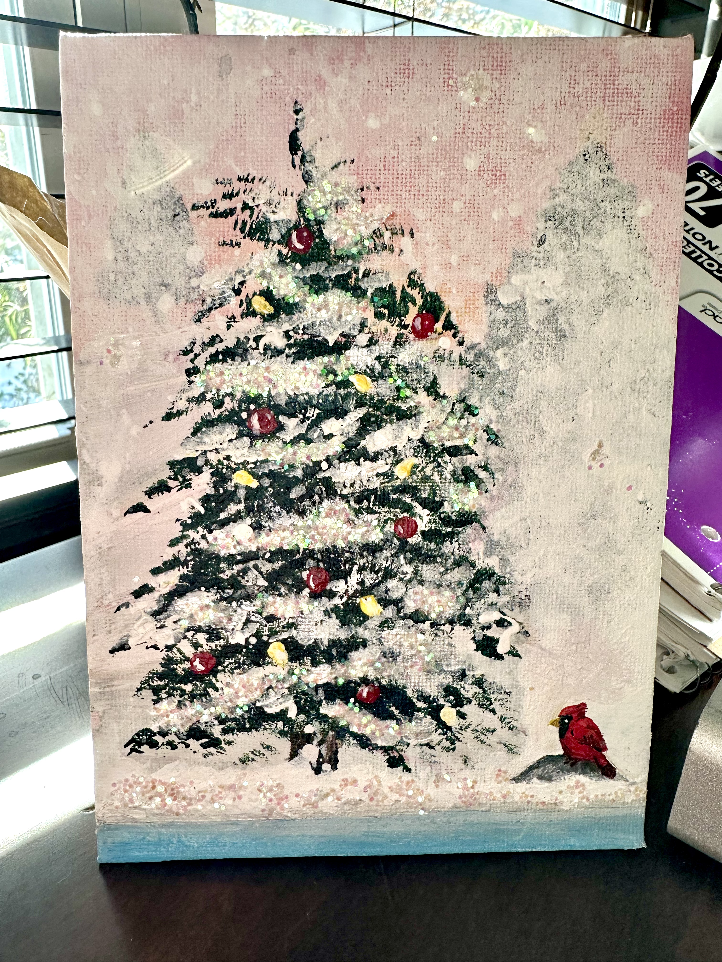 A painting of a decorated Christmas tree with red and yellow ornaments, set against a pink background, with a red cardinal bird sitting on the snow at the bottom right corner.