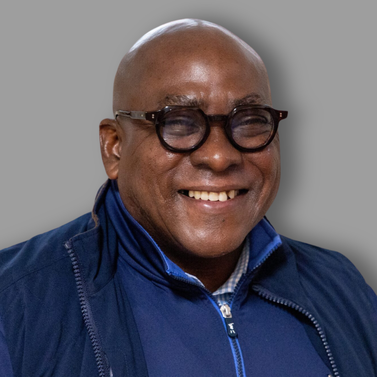 A smiling man with dark skin, wearing glasses, a blue jacket, and a checkered shirt, facing the camera against a gray background.